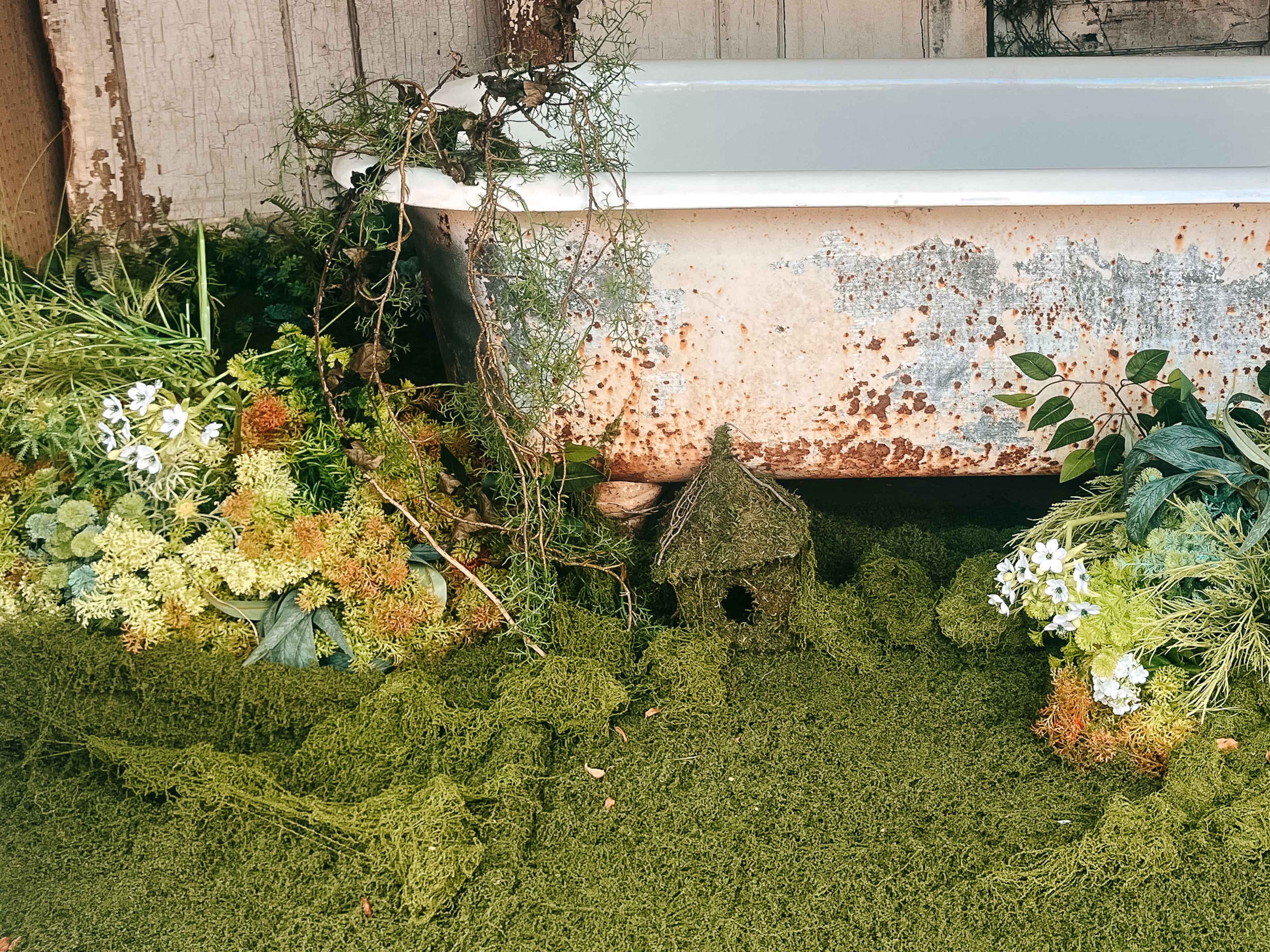 An old, rusty bathtub sits among lush greenery and decorative plants, with a small birdhouse partially obscured by moss and foliage.