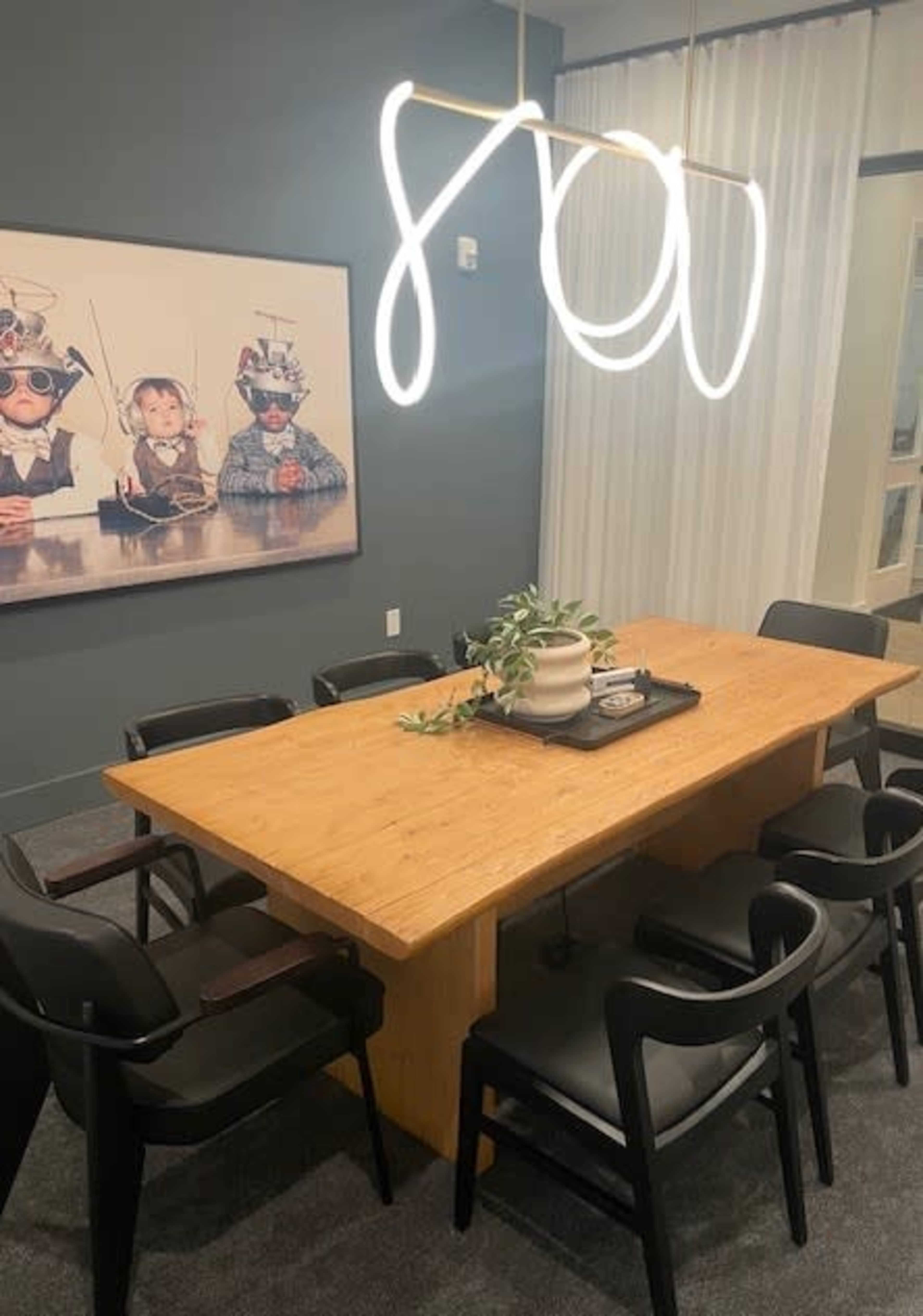 A modern dining area featuring a wooden table surrounded by black chairs, with a unique light fixture overhead and a wall art piece depicting two children in playful costumes.