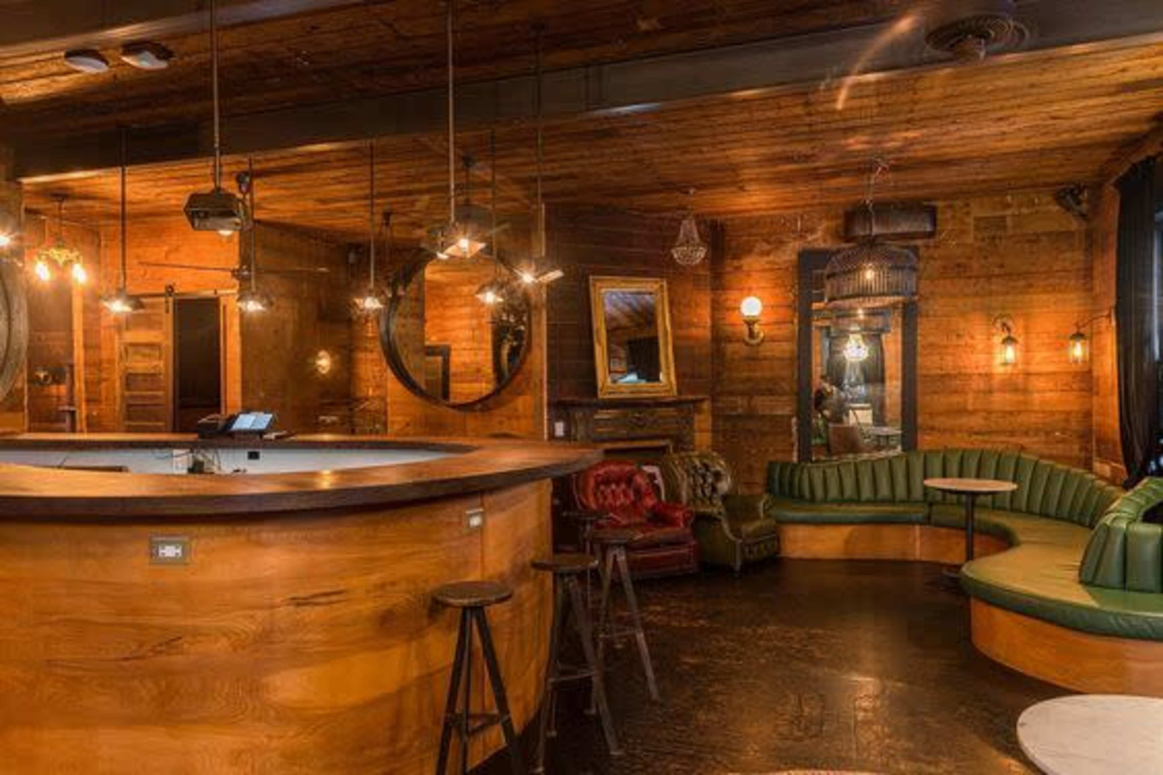 A dimly lit bar interior featuring wooden walls, circular mirrors, and a mix of seating arrangements including a curved green leather bench and red armchairs.