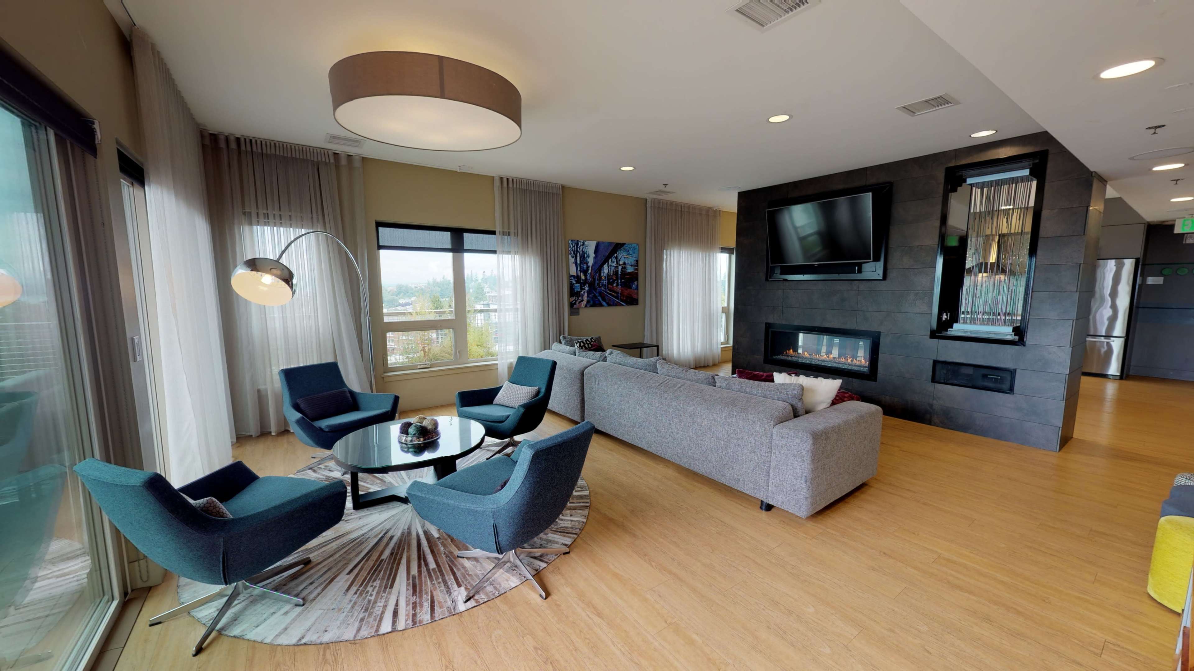 A modern living area features a gray sofa, glass coffee table, and four blue chairs, complemented by a fireplace and large windows with natural light.