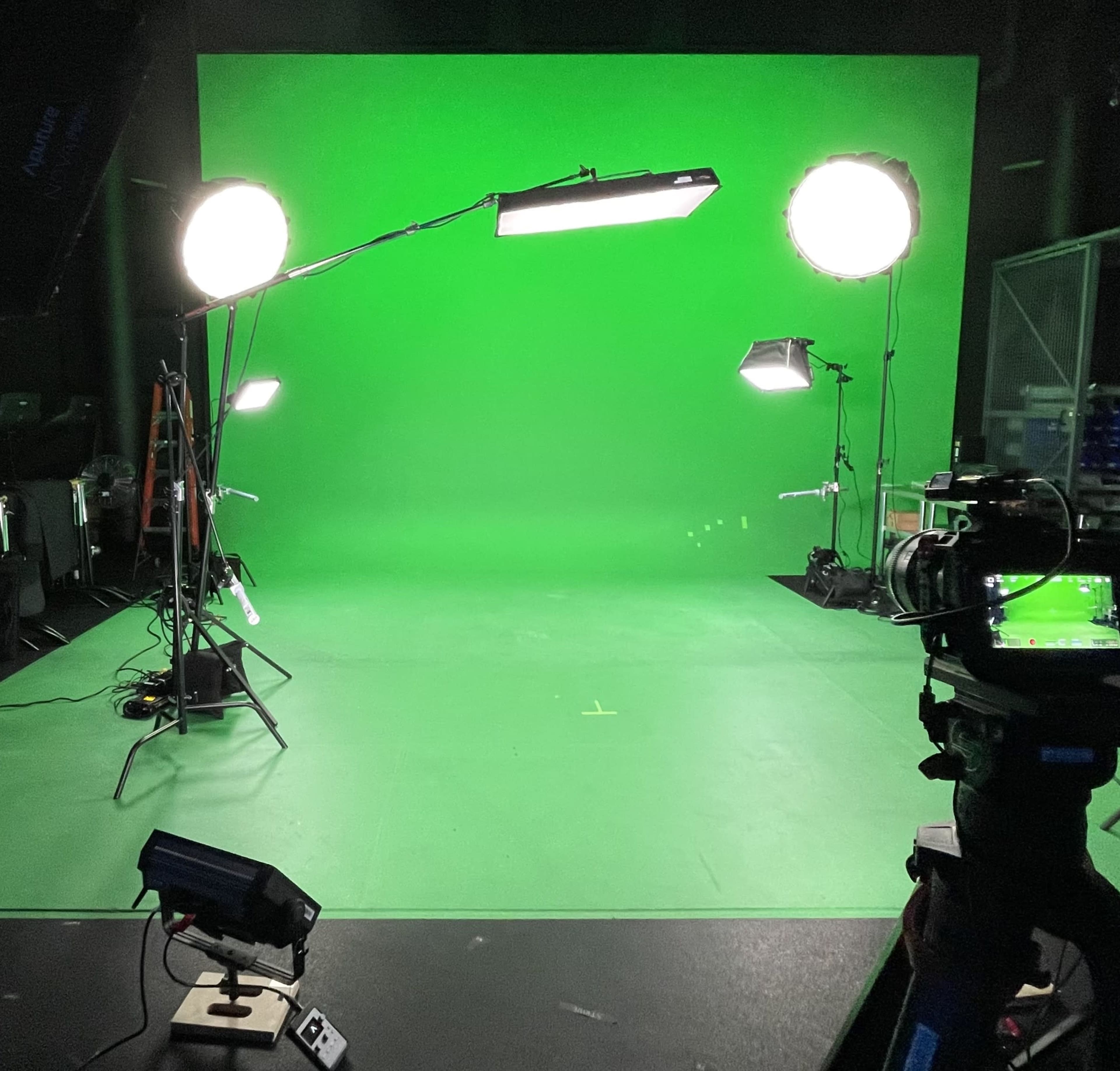 A photography studio features a large green screen surrounded by multiple lights and camera equipment.