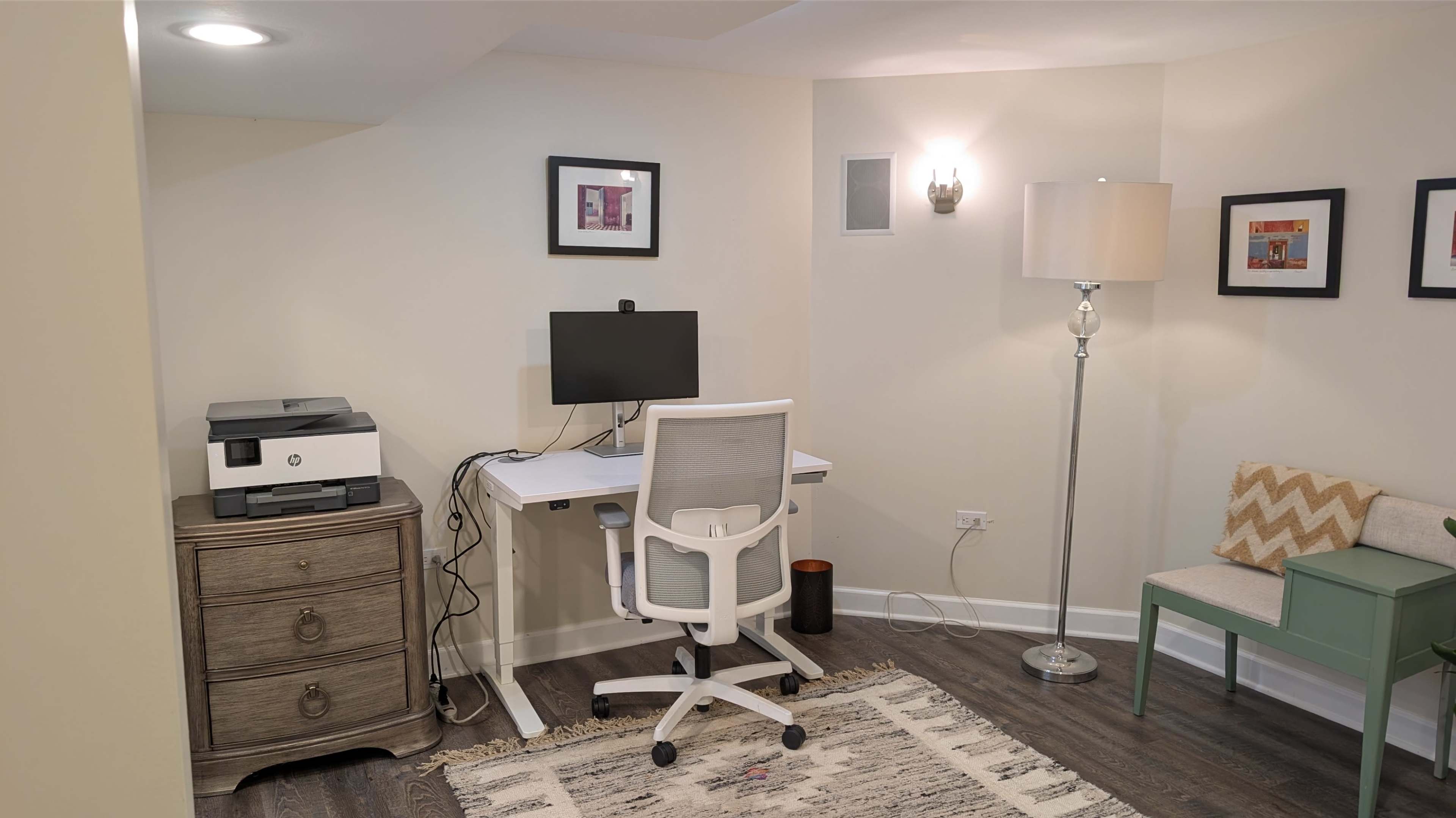 The image shows a home office setup featuring a desk with a computer monitor, a chair, a printer, and a lamp, along with a patterned cushion on a small bench and a decorative rug on the floor.