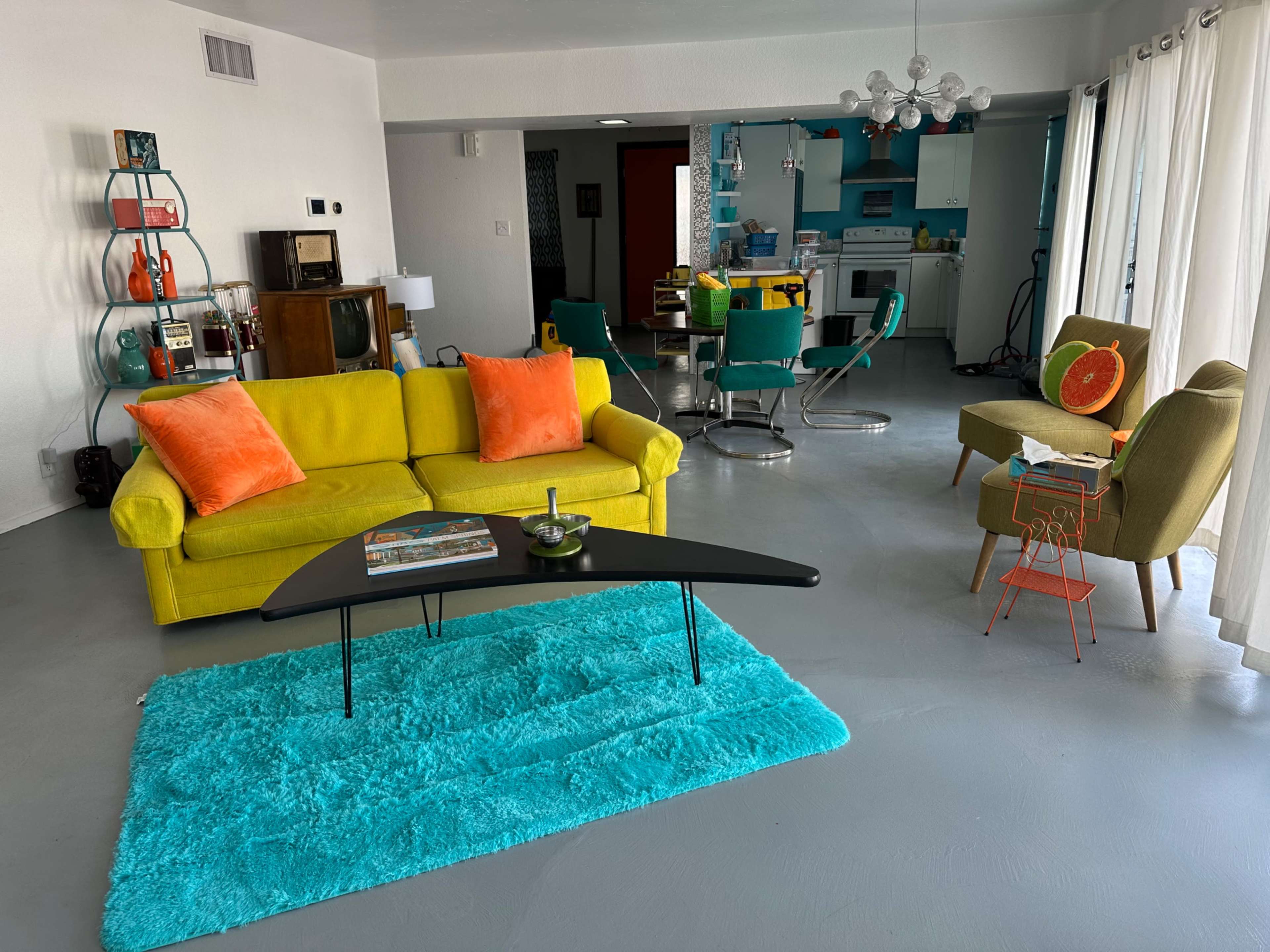 The image shows a brightly colored living space featuring a yellow sofa, a black coffee table, a turquoise rug, and a dining area with retro-style chairs and a kitchen in the background.