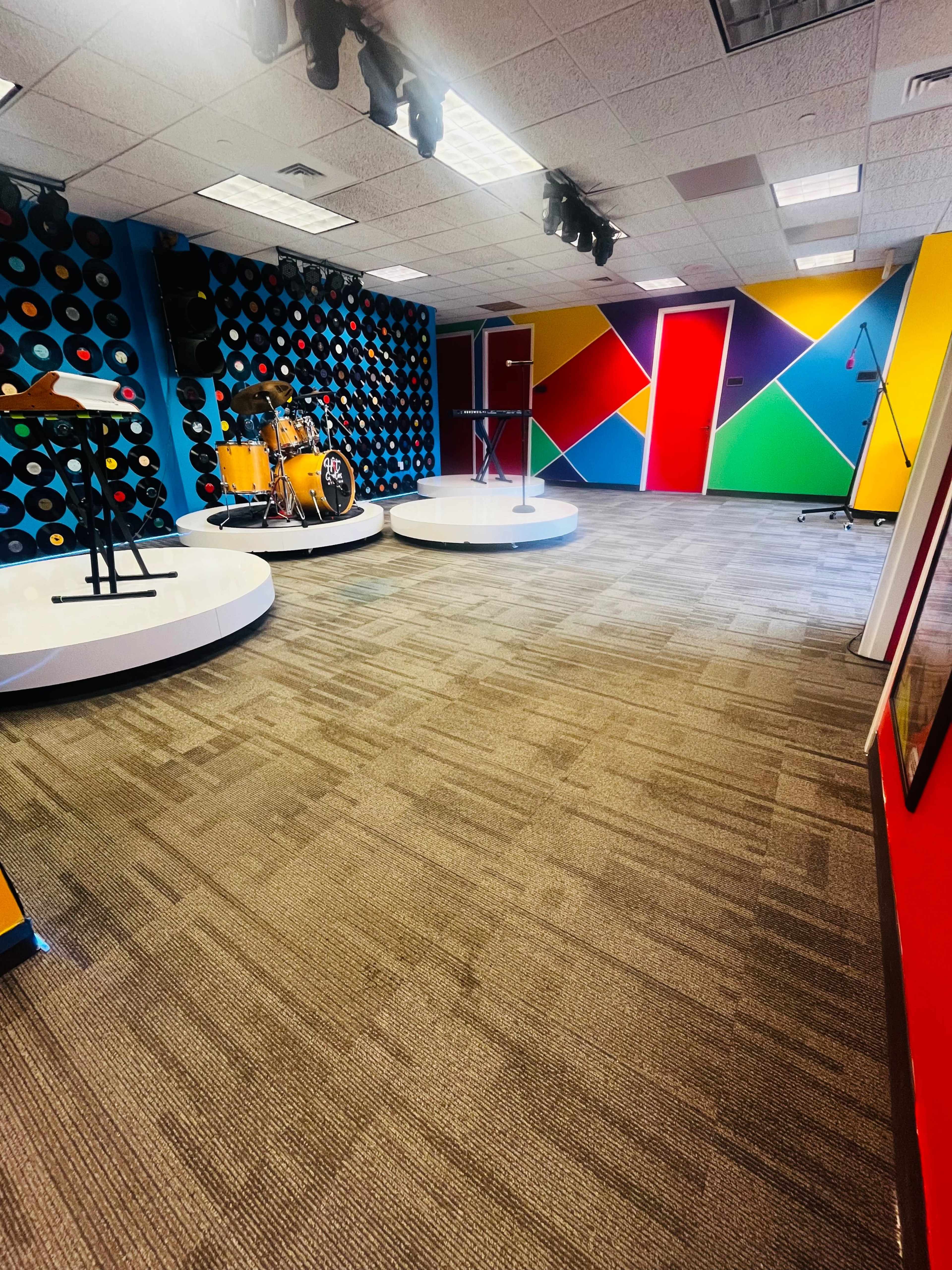 A music practice room featuring a drum set, music stands, and colorful geometric wall decorations with a patterned backdrop of vinyl records.