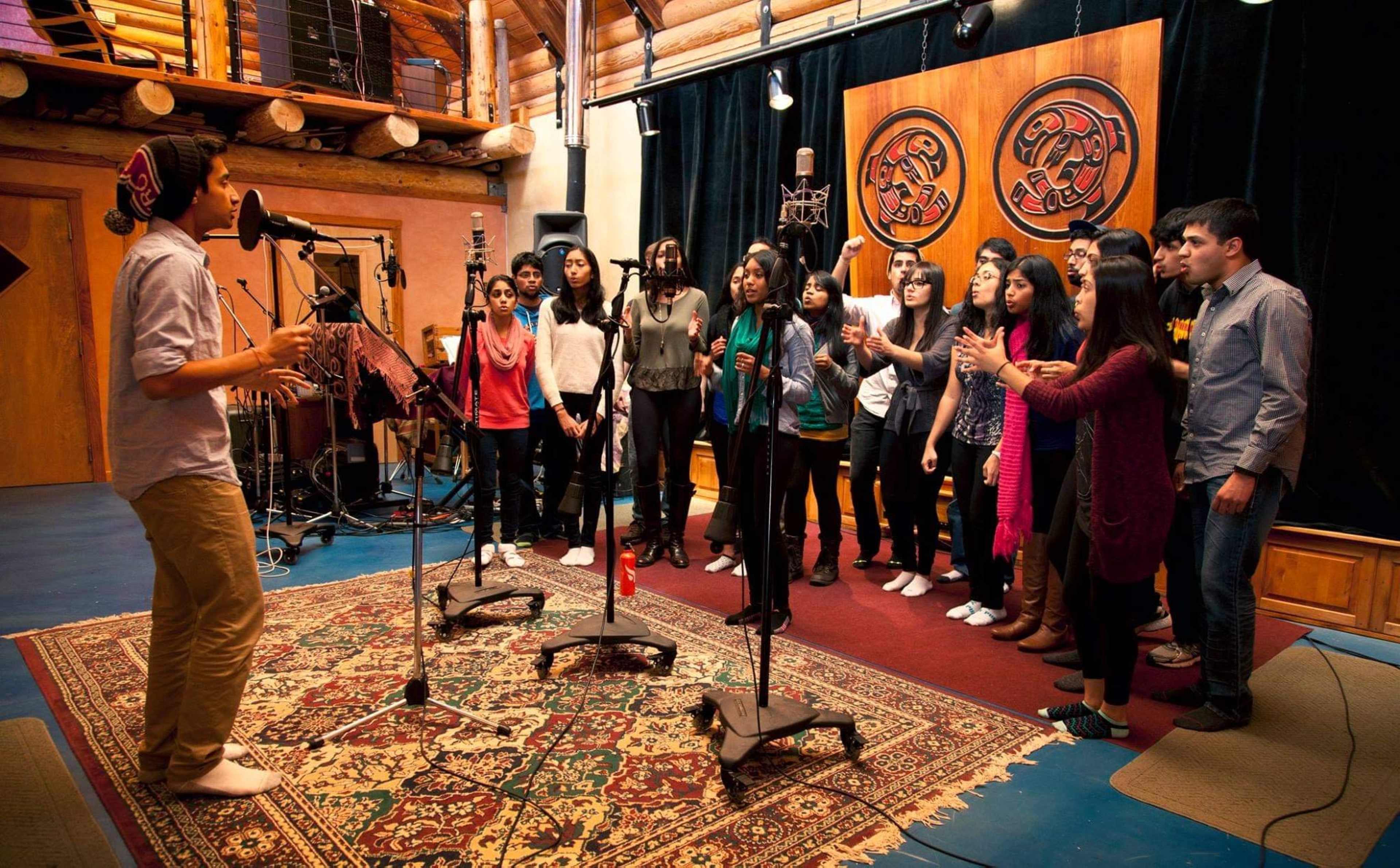 A group of young singers stands in a recording studio while a male vocalist performs in front of them.