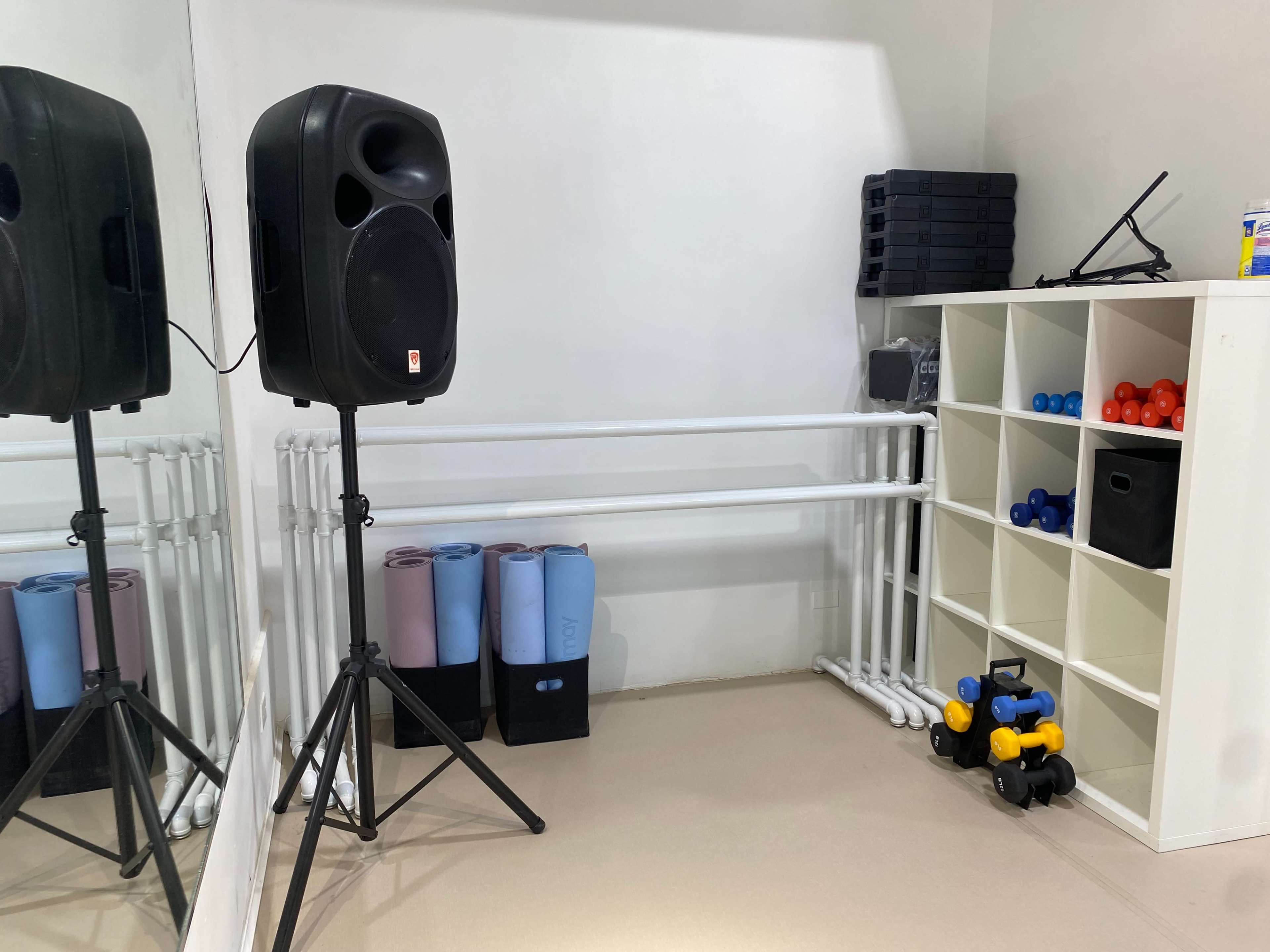 A fitness room equipped with a speaker, exercise mats, a shelving unit with colored weights, and a set of dumbbells on the floor.