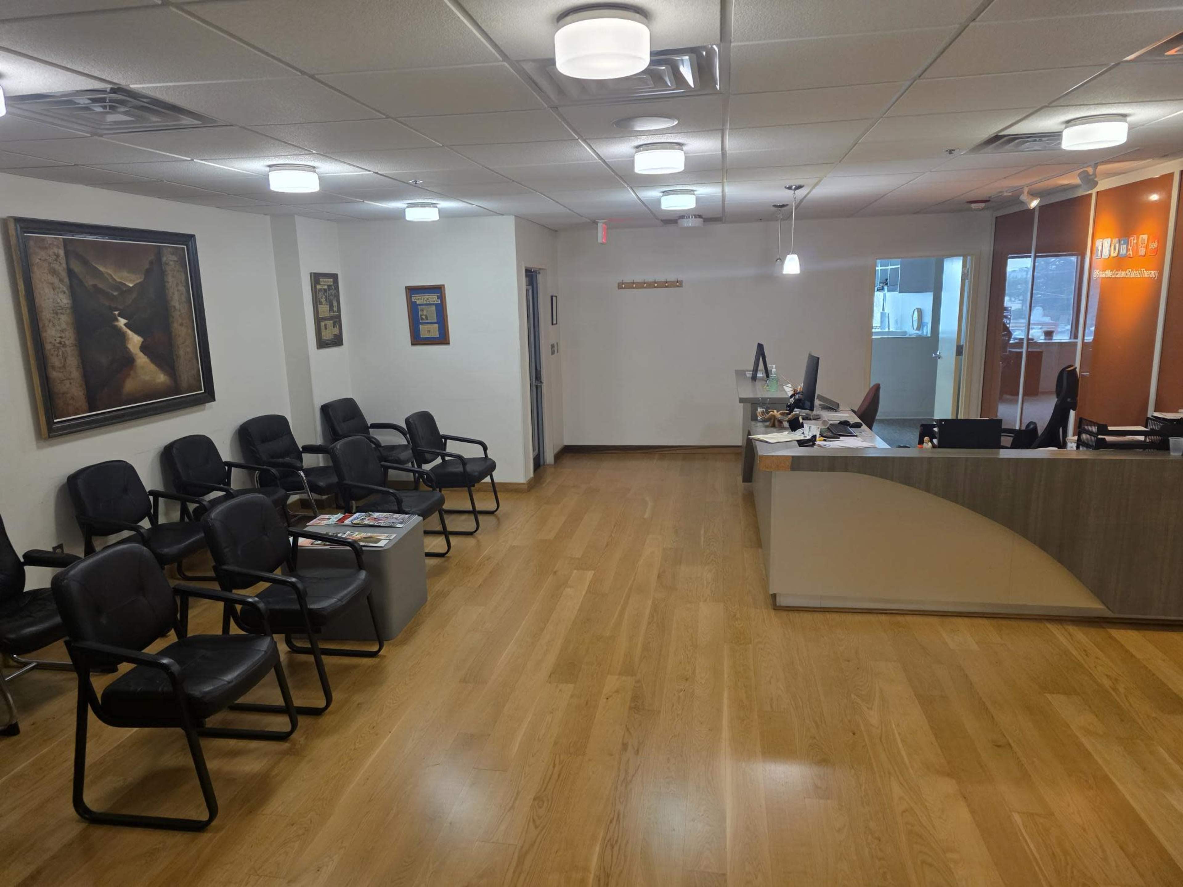 The image shows a modern waiting room with black chairs, a reception desk, and hardwood floors.