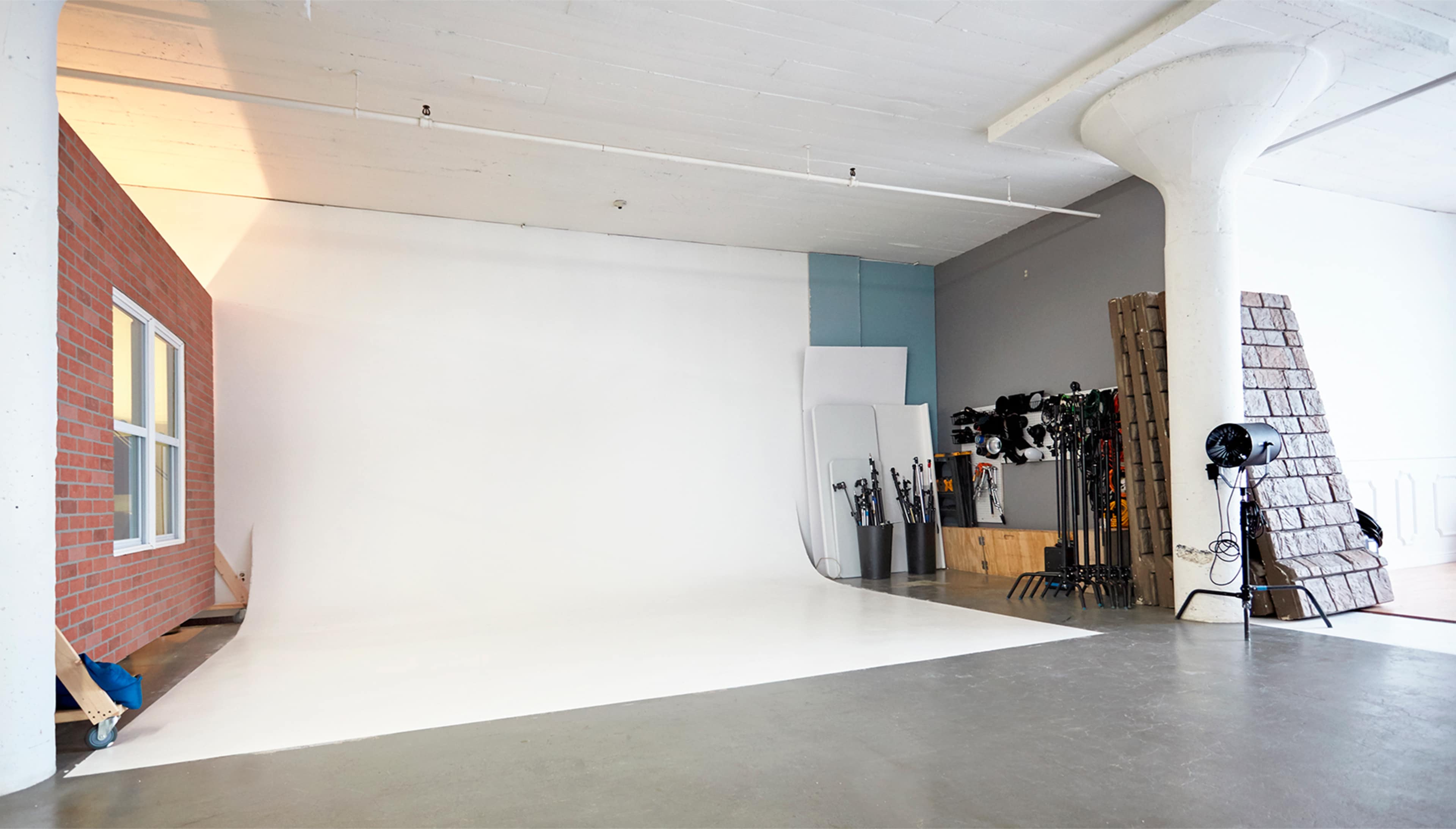 The image shows a spacious photography studio with a white backdrop, a brick wall, and various lighting equipment arranged against the walls.