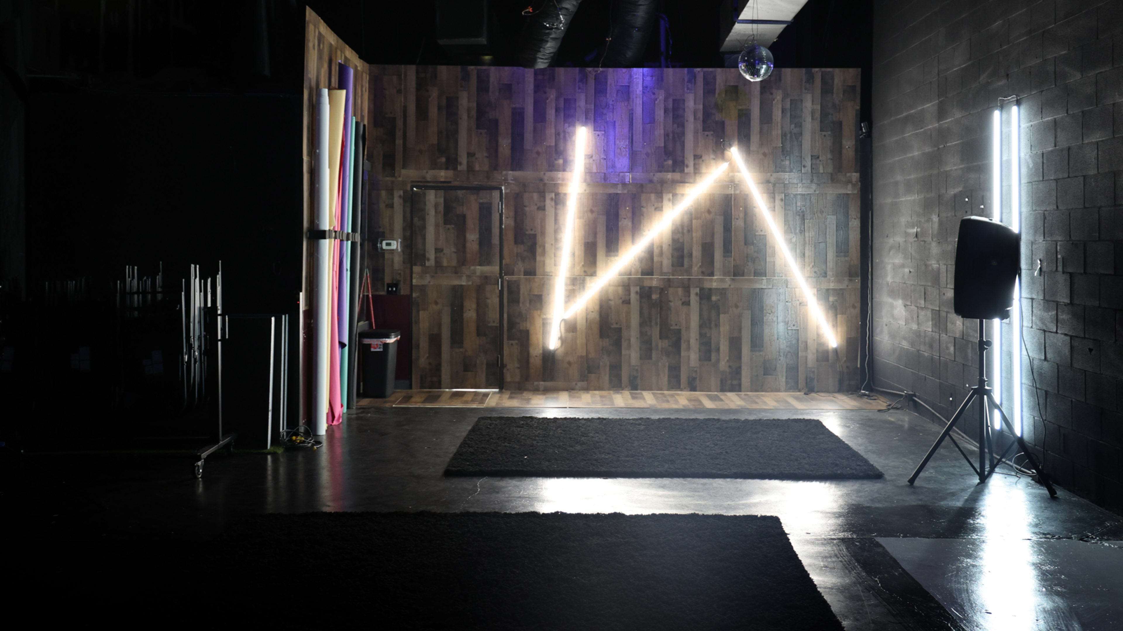 A dimly lit room with wooden walls, featuring neon lights arranged in a "W" shape and black carpets on the floor.