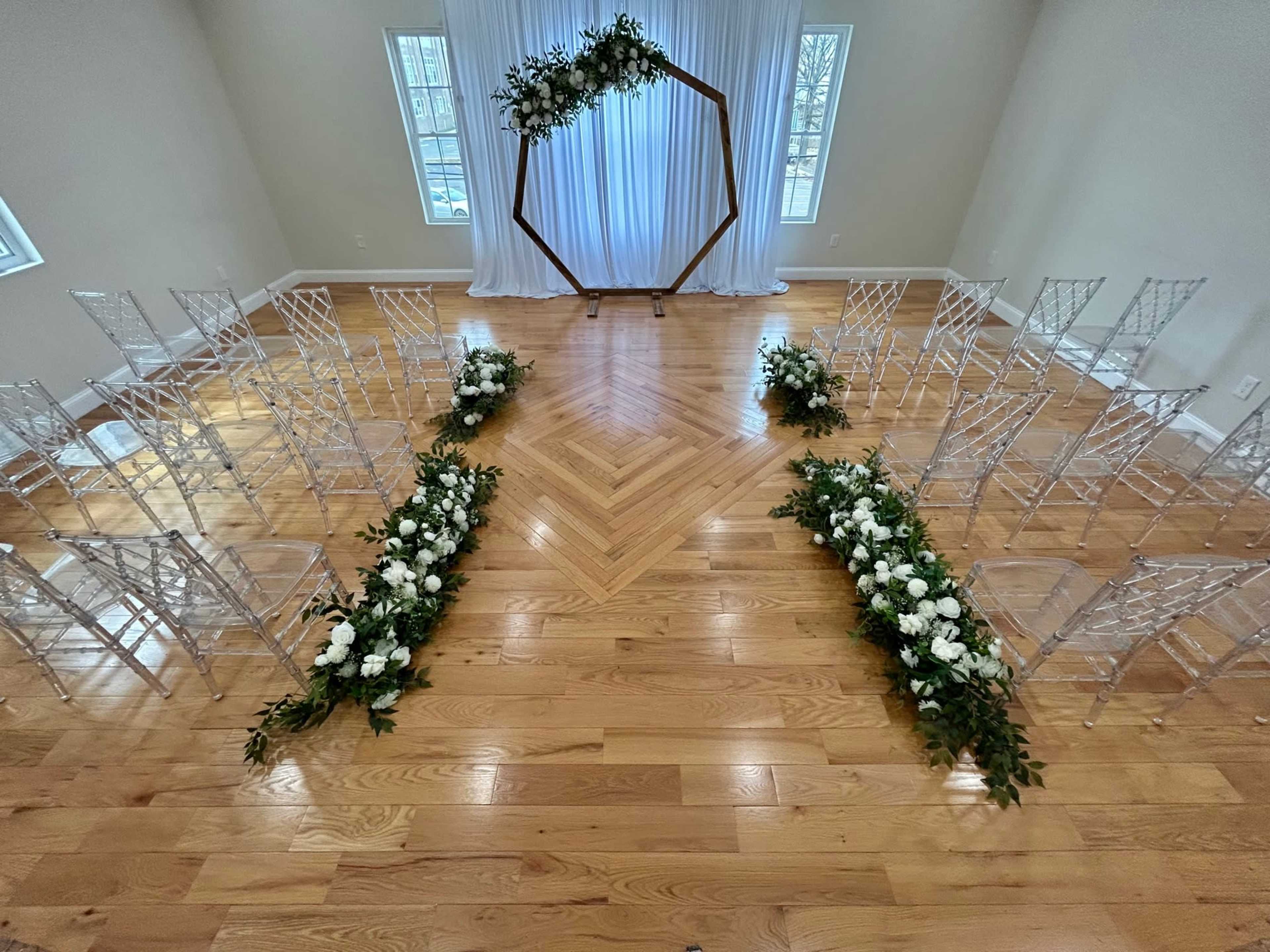 The image shows a wedding ceremony setup featuring clear chairs arranged around a floral-accented aisle, leading to a hexagonal wooden arch draped with fabric.