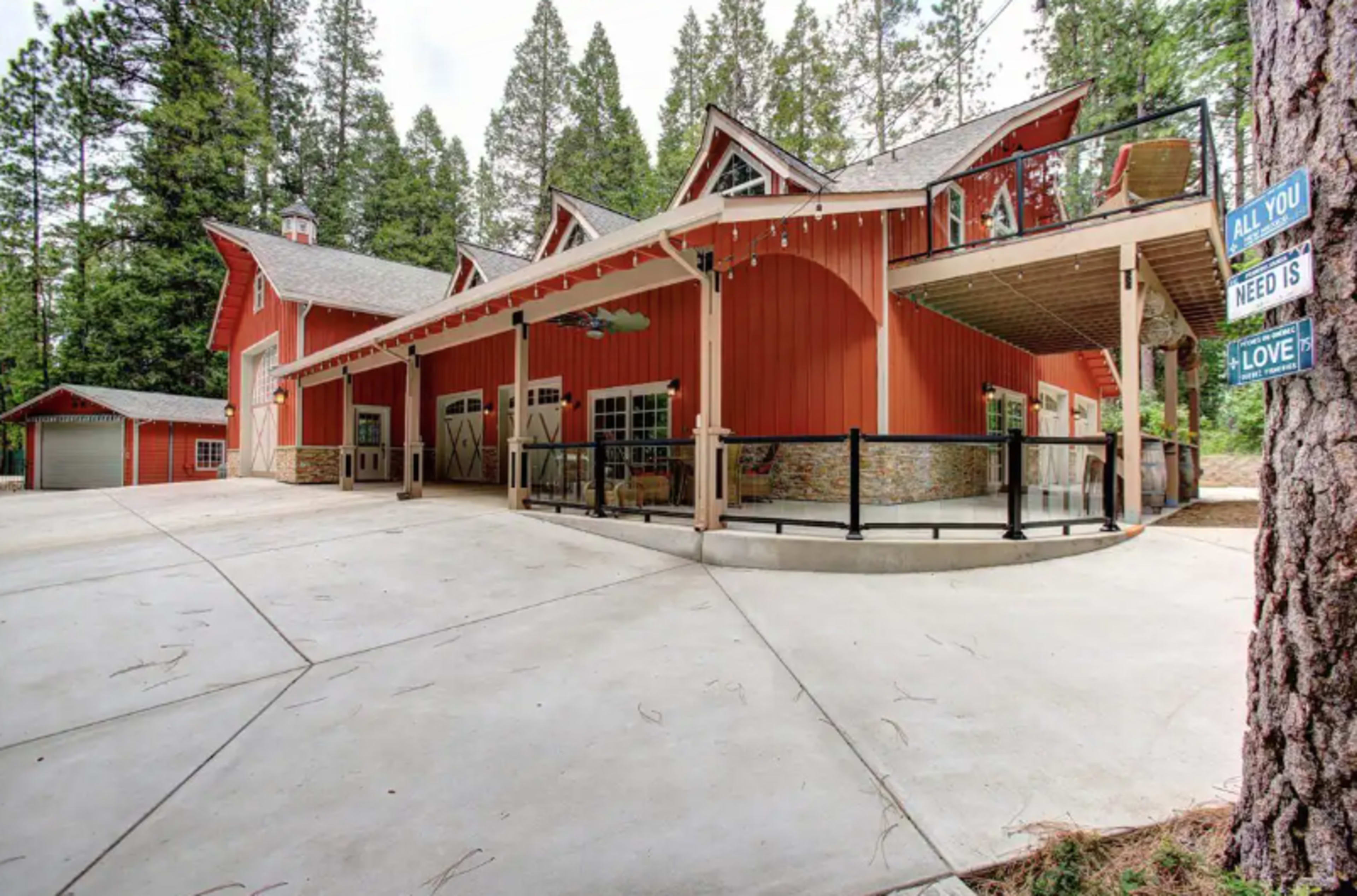 A red and gray multi-level house with a wraparound porch, surrounded by tall green trees.
