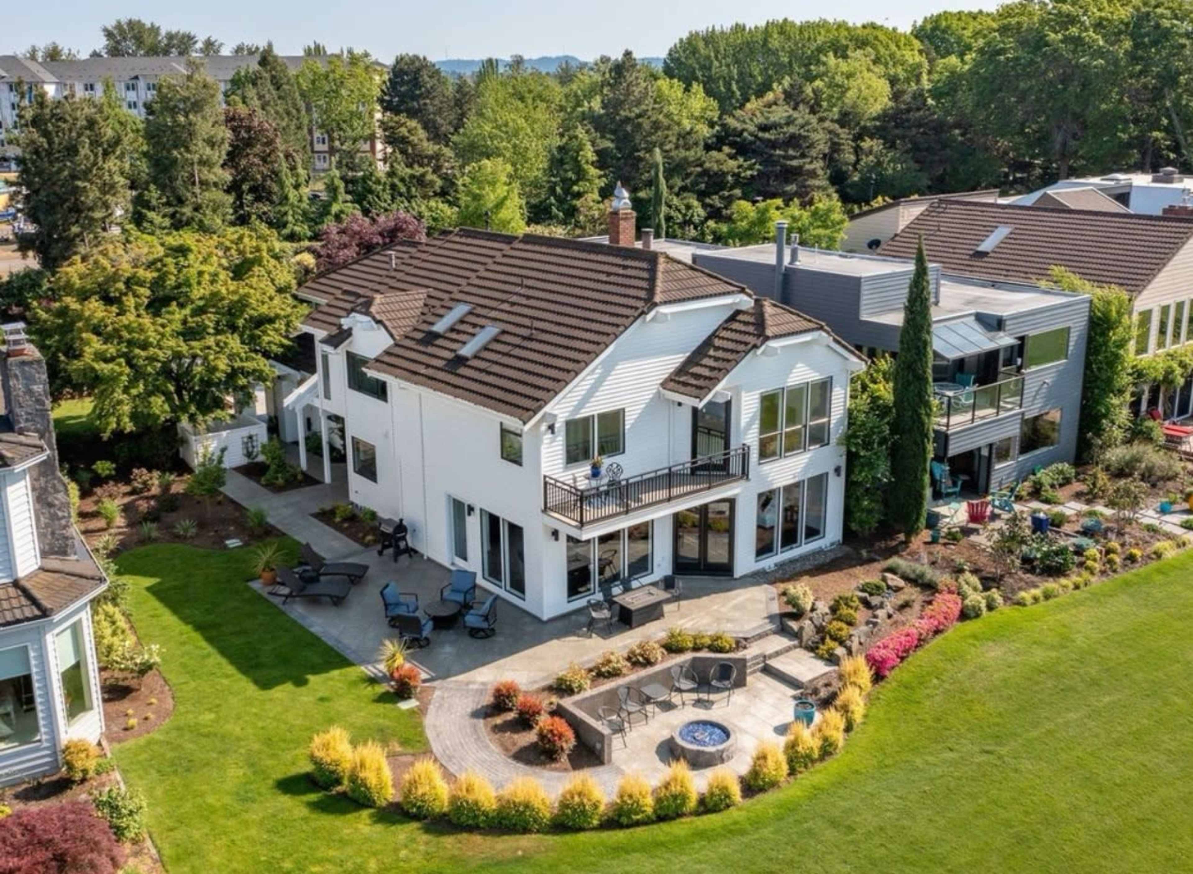 The image shows a large, two-story house with a patio and landscaped garden, surrounded by trees and other homes.