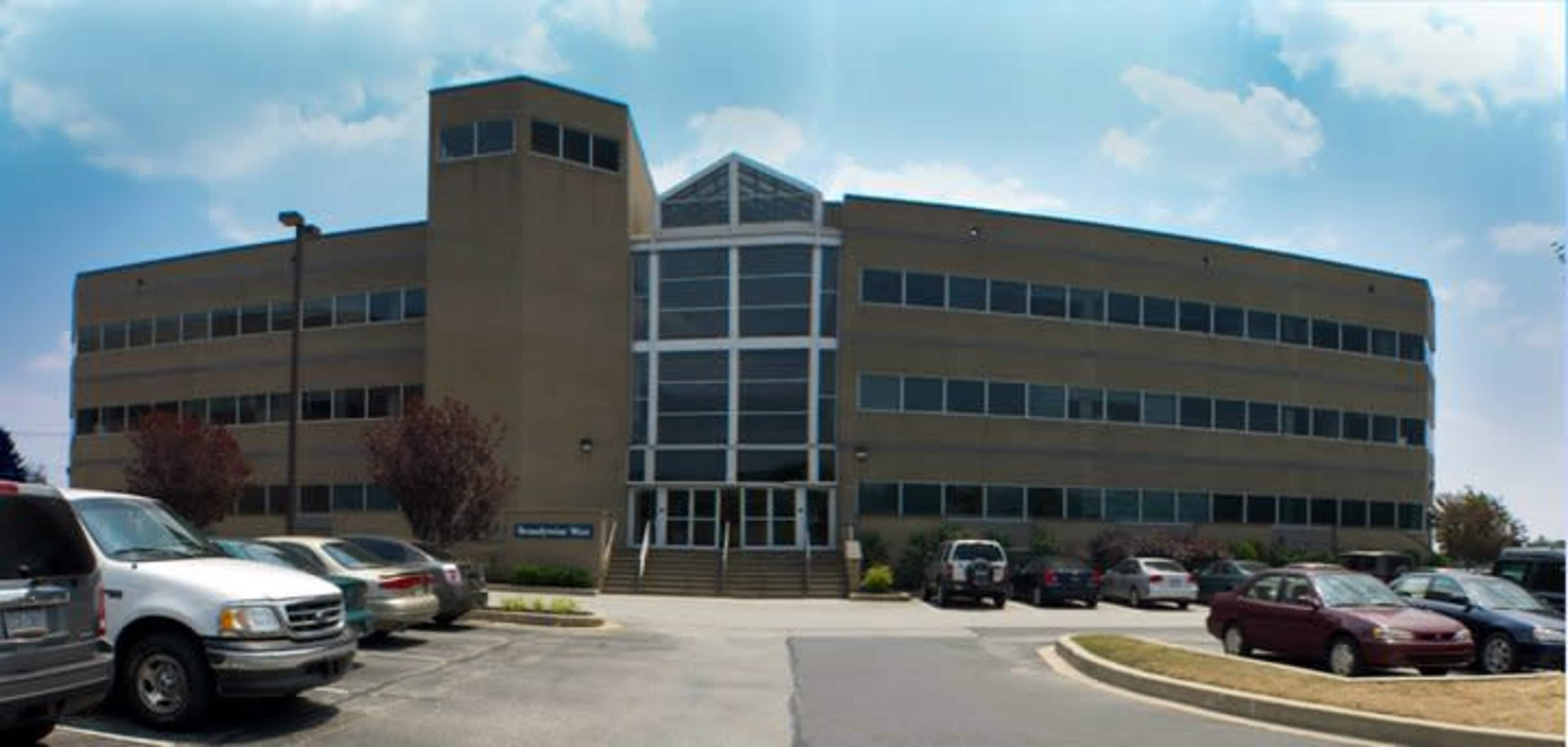 A modern, three-story office building features large windows and is surrounded by a parking lot filled with several vehicles.