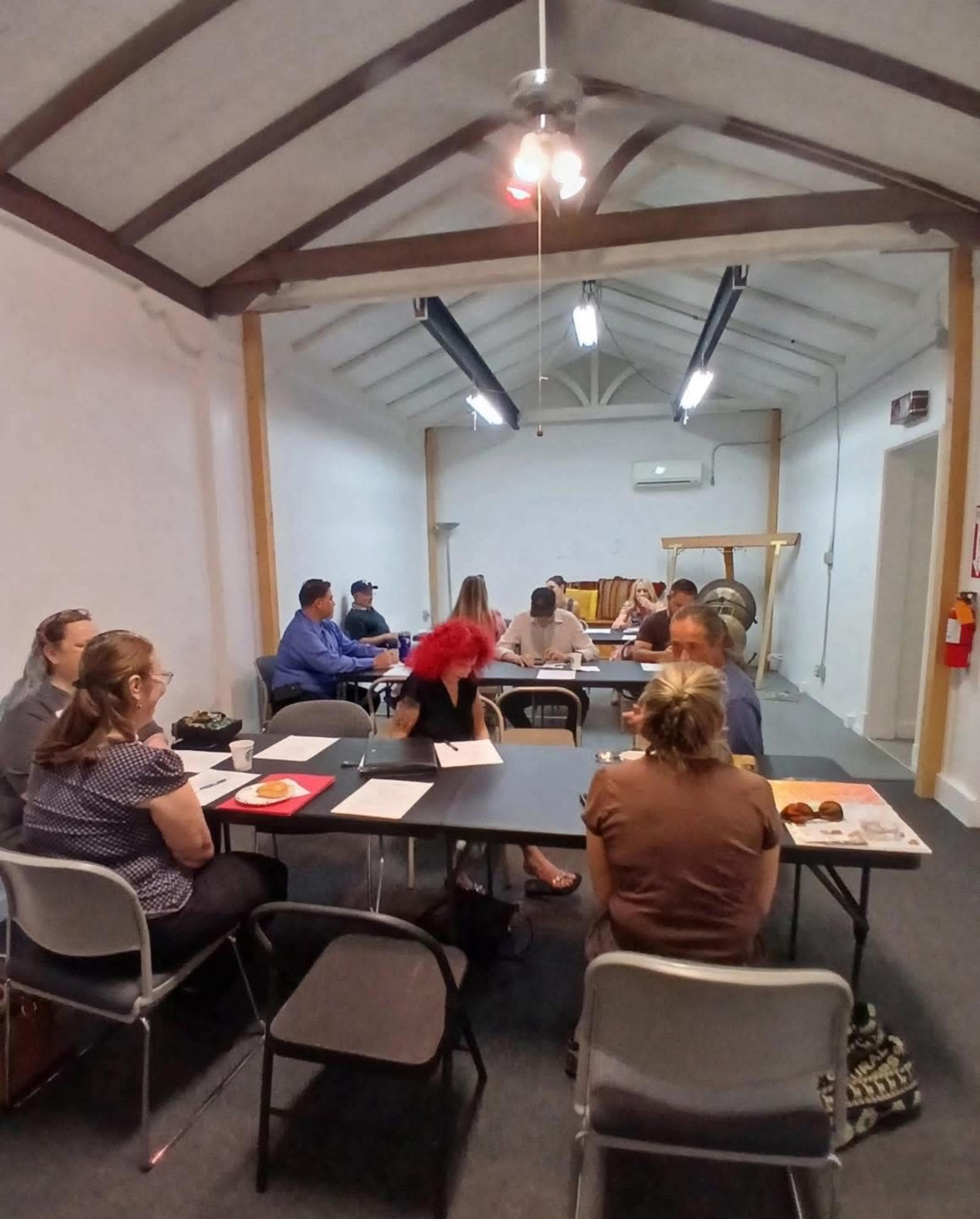 A group of people are seated around tables in a meeting room with a high ceiling and exposed beams.