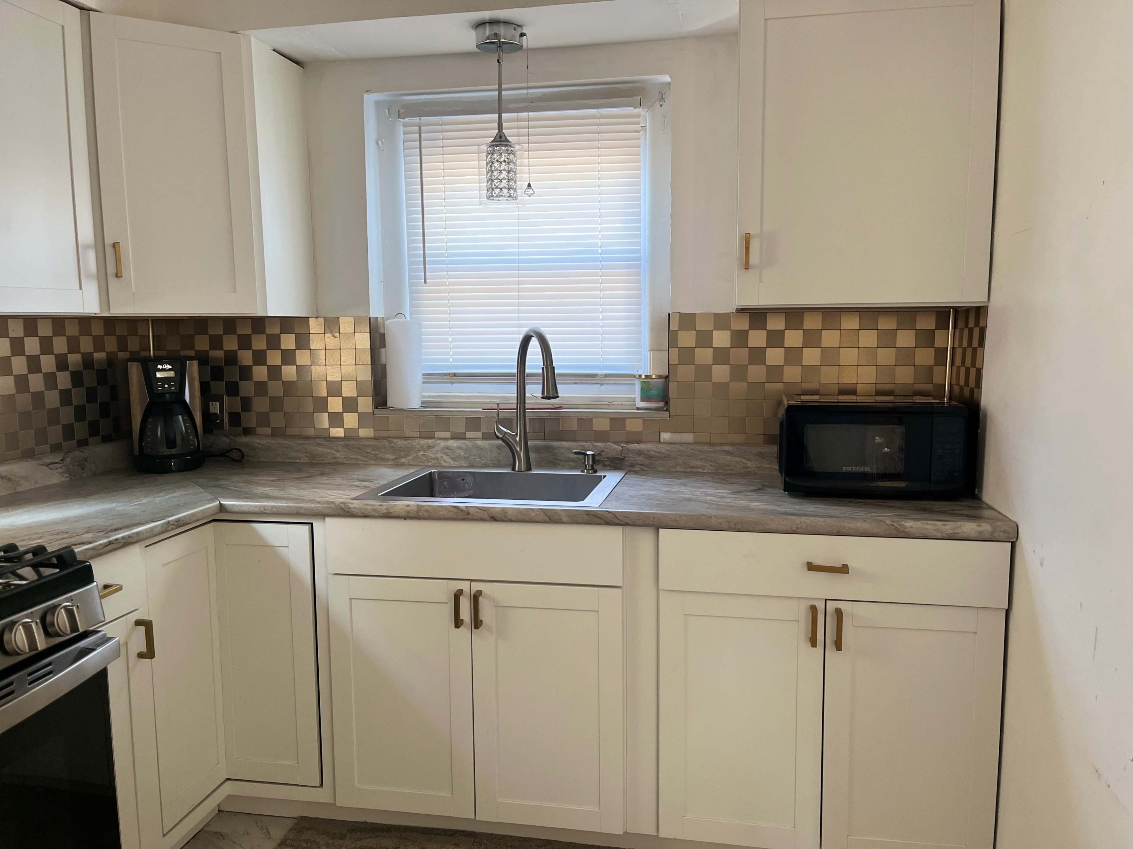 The image shows a modern kitchen with white cabinets, a stainless steel sink, a stove, a coffee maker, and a microwave, featuring a window with blinds.