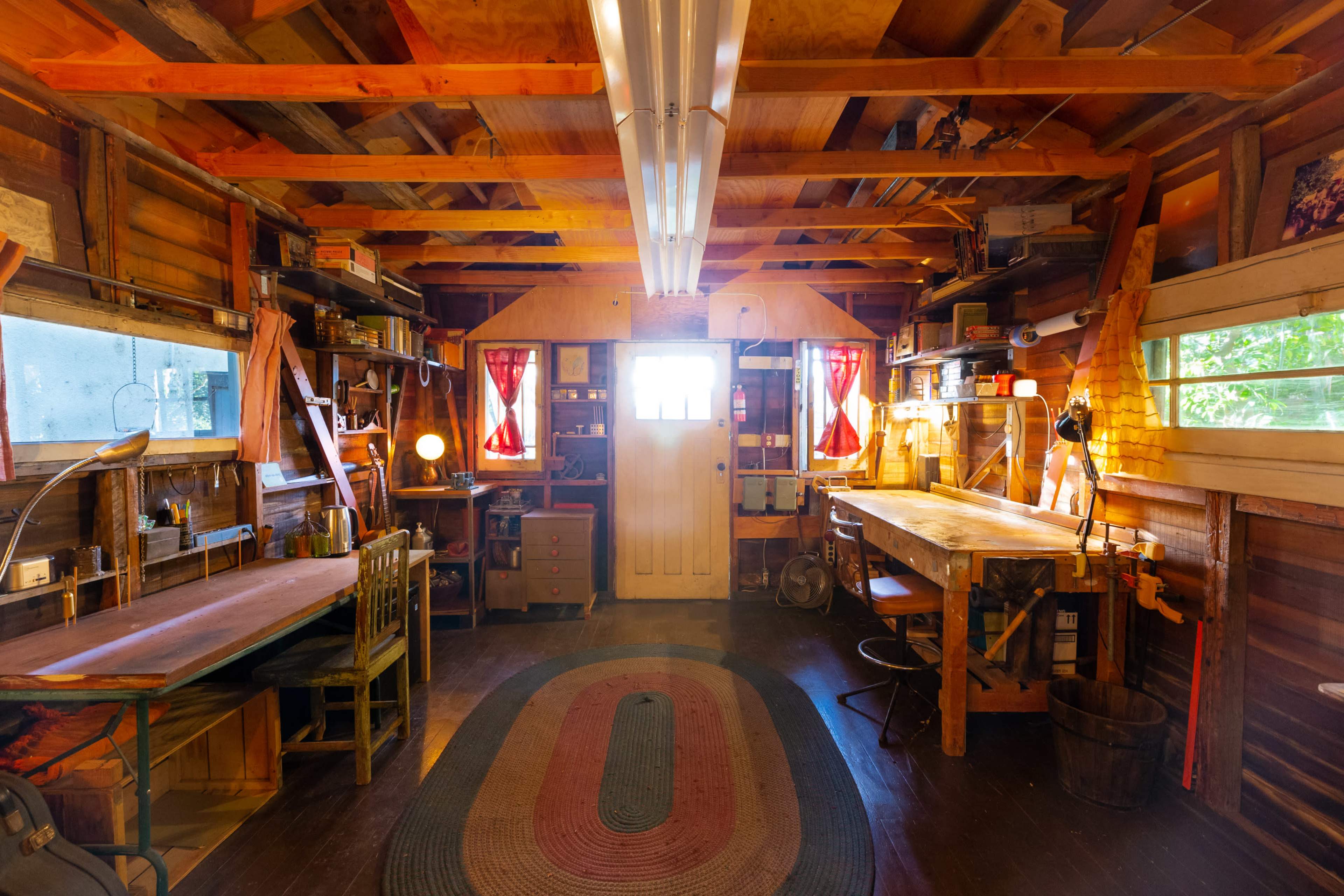 A wooden workshop with two large workbenches, shelves filled with tools and materials, and a door leading outside.