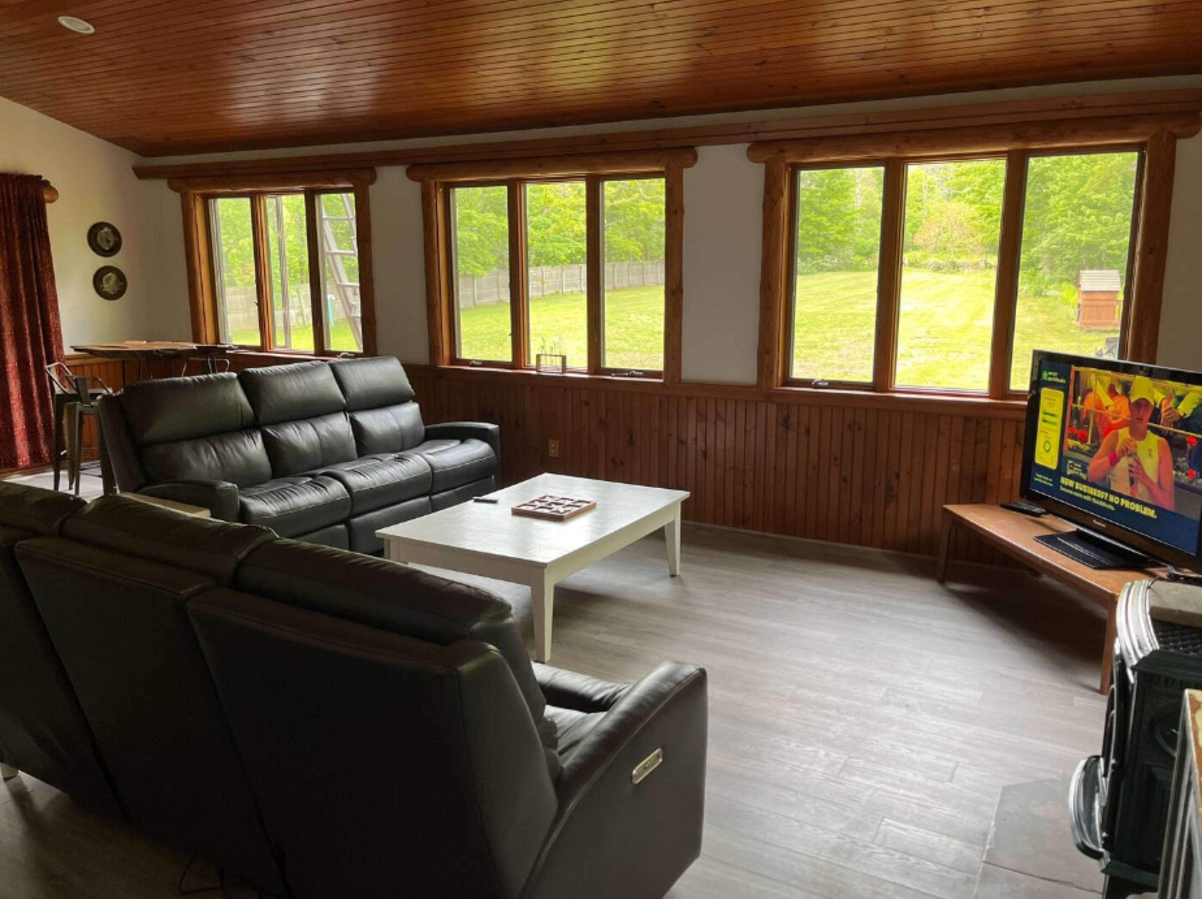 A sunlit living room with a black leather couch, a white coffee table, and a television, surrounded by large windows and wooden walls.