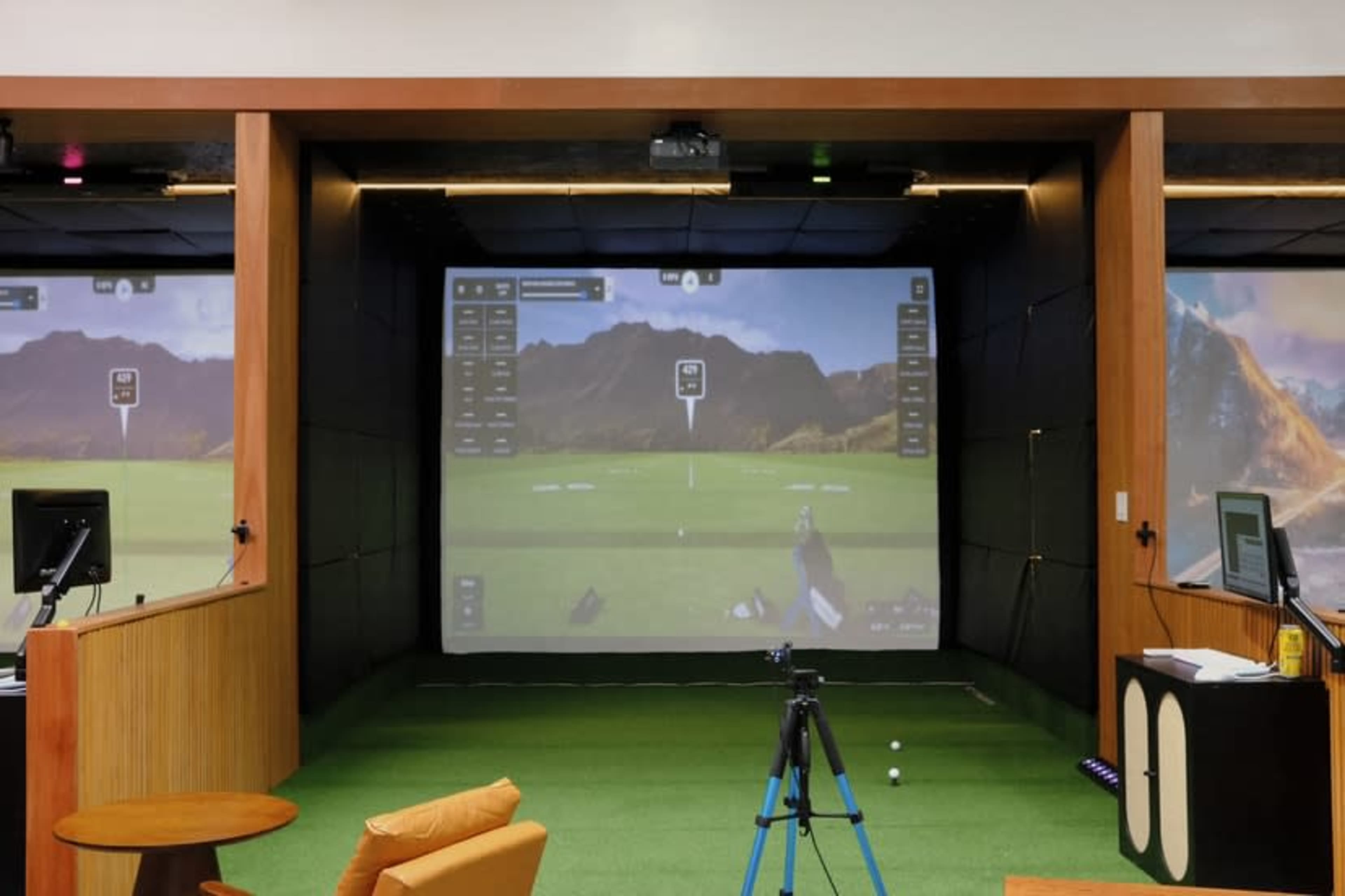 The image shows a modern golf simulator setup featuring a large screen displaying a virtual golf course, with artificial grass flooring and a tripod in the foreground.