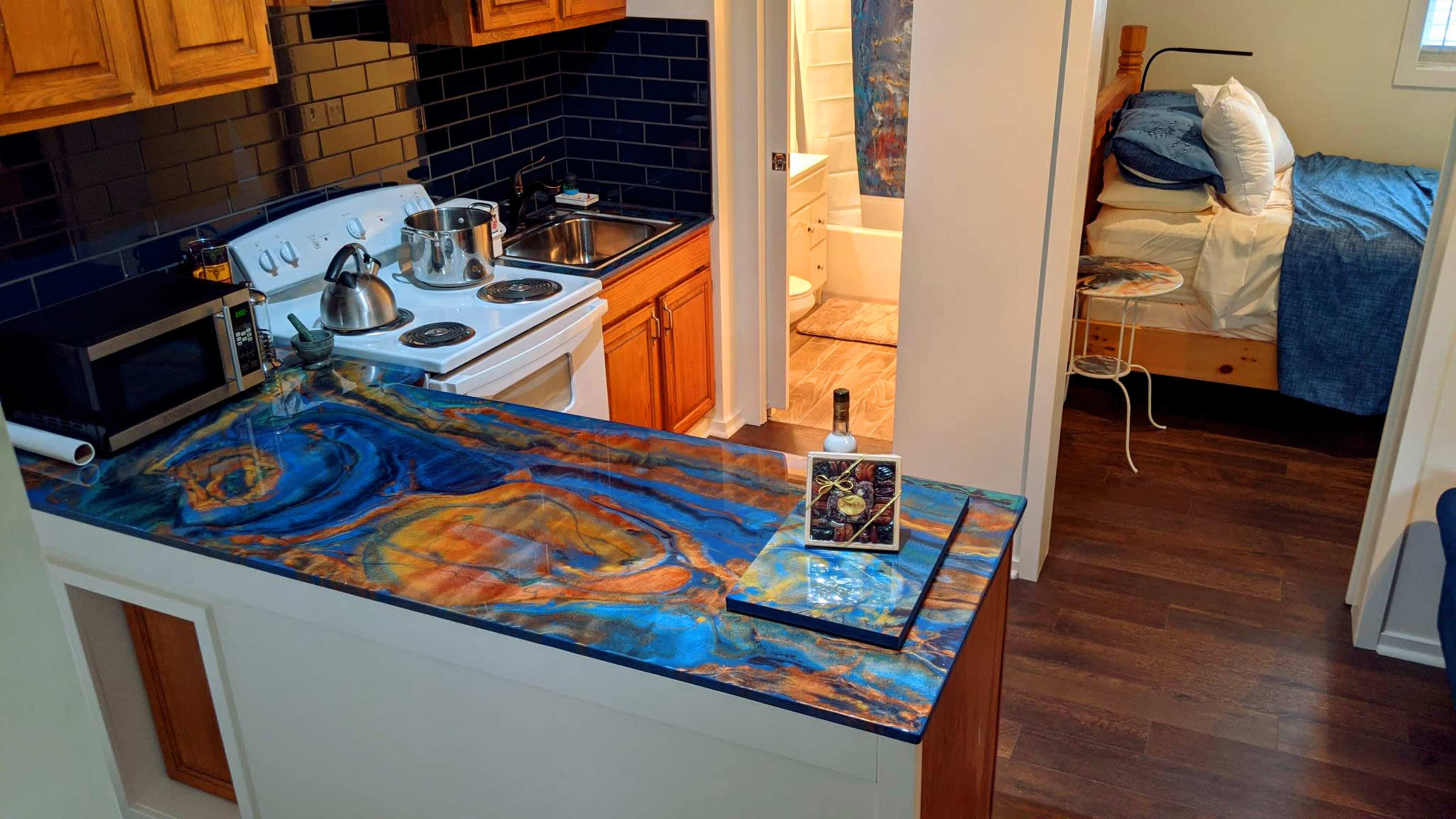 The image shows a kitchen with a blue and orange marbled countertop, a sink, stove, and microwave, leading into a small living area with a bed and bathroom visible.