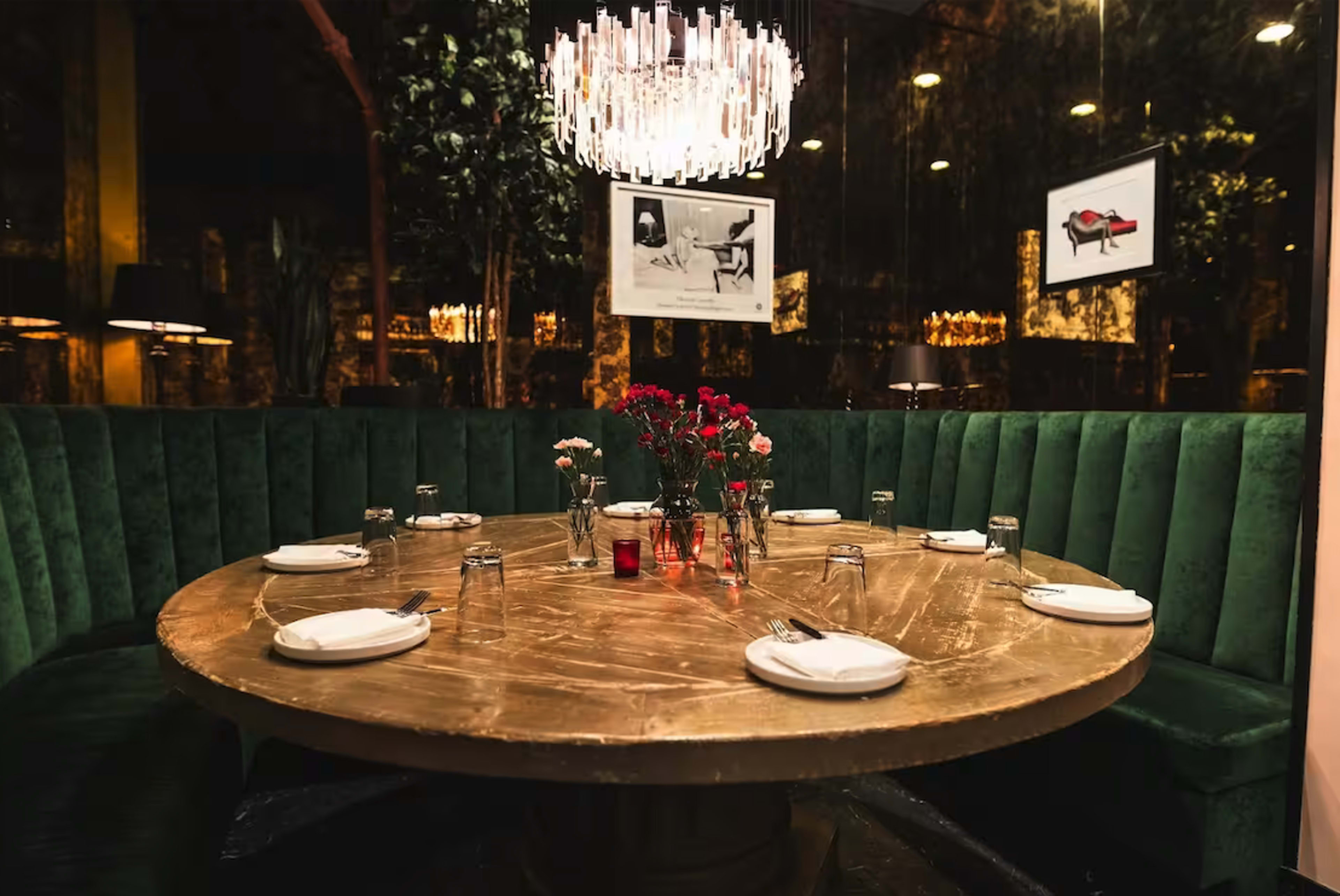 A round table with white plates is set in a green upholstered booth, surrounded by decorative plants and illuminated by a chandelier.