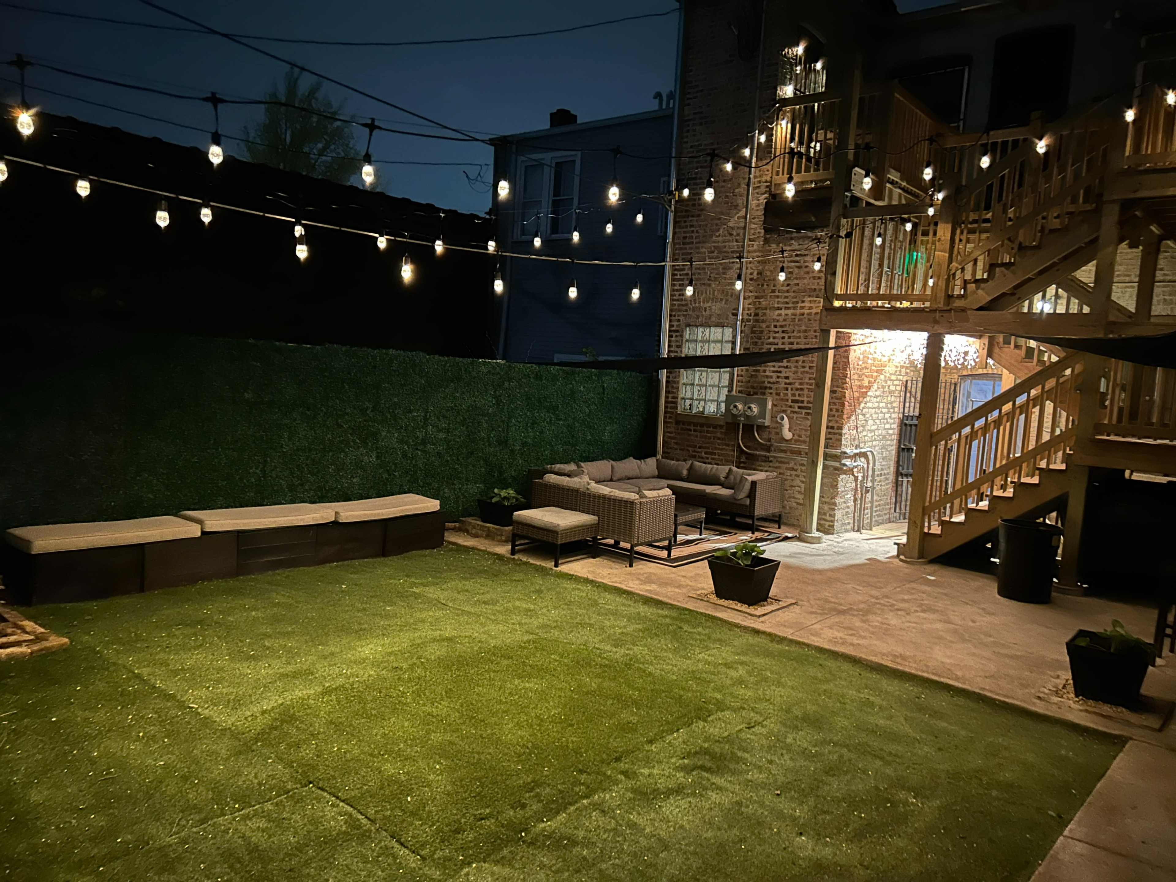 A dimly lit backyard featuring artificial turf, a seating area with gray wicker furniture, and string lights overhead.