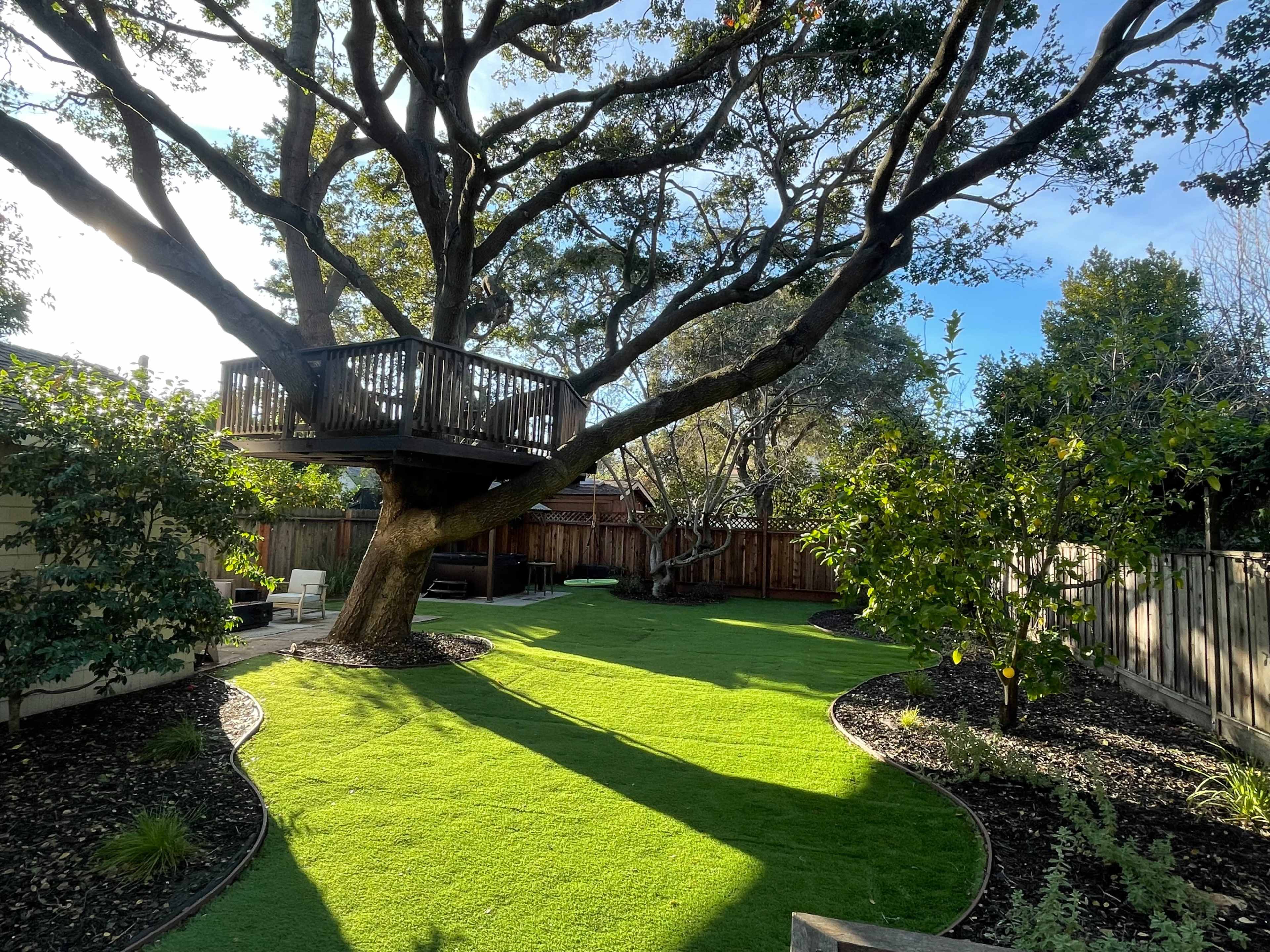 A wooden treehouse is built high in a large tree, surrounded by a well-manicured lawn and landscaped garden.