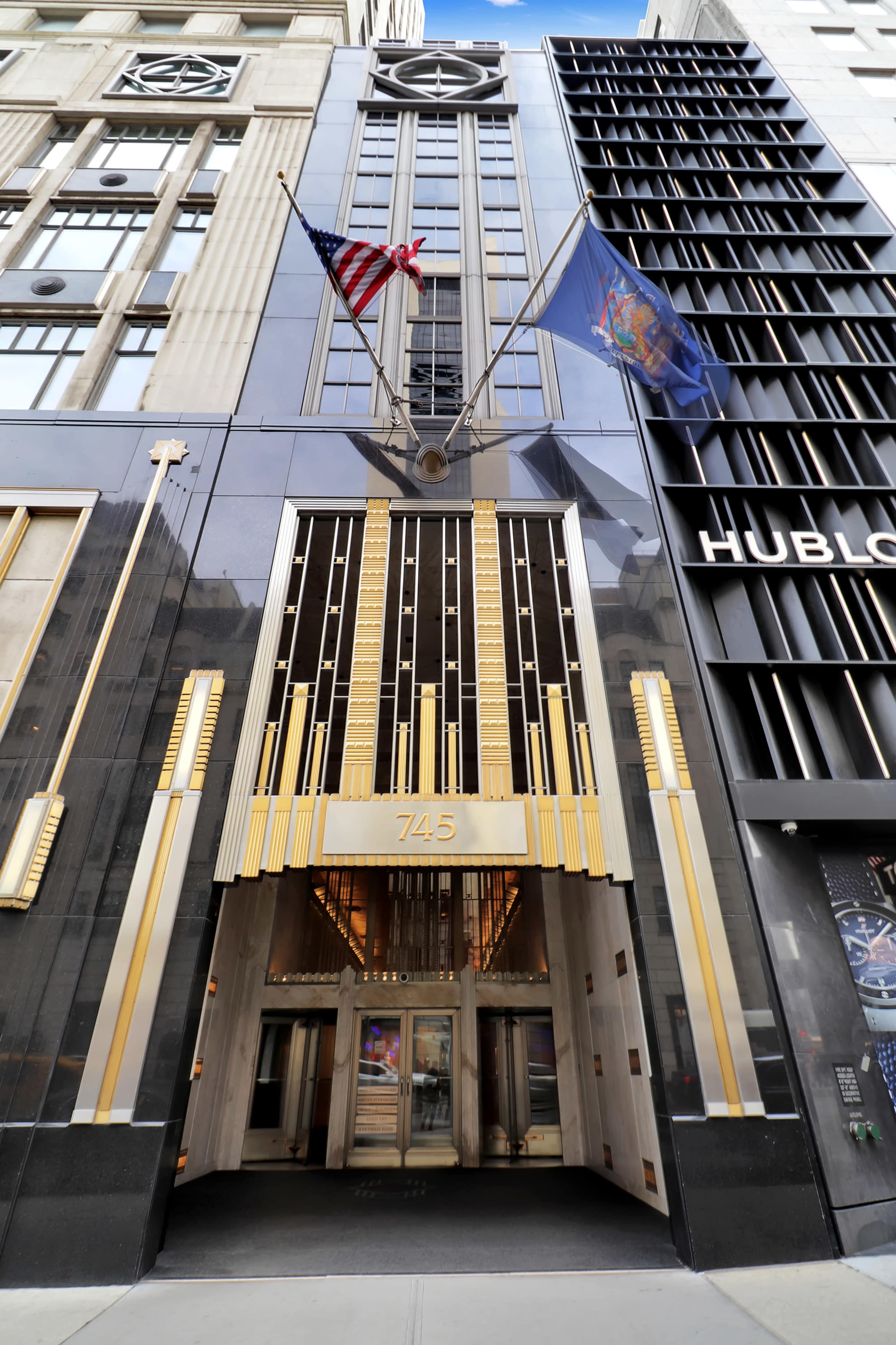 The image shows the entrance of a tall building featuring an Art Deco design with an American flag and a state flag displayed above the entrance, which is marked with the number "745."
