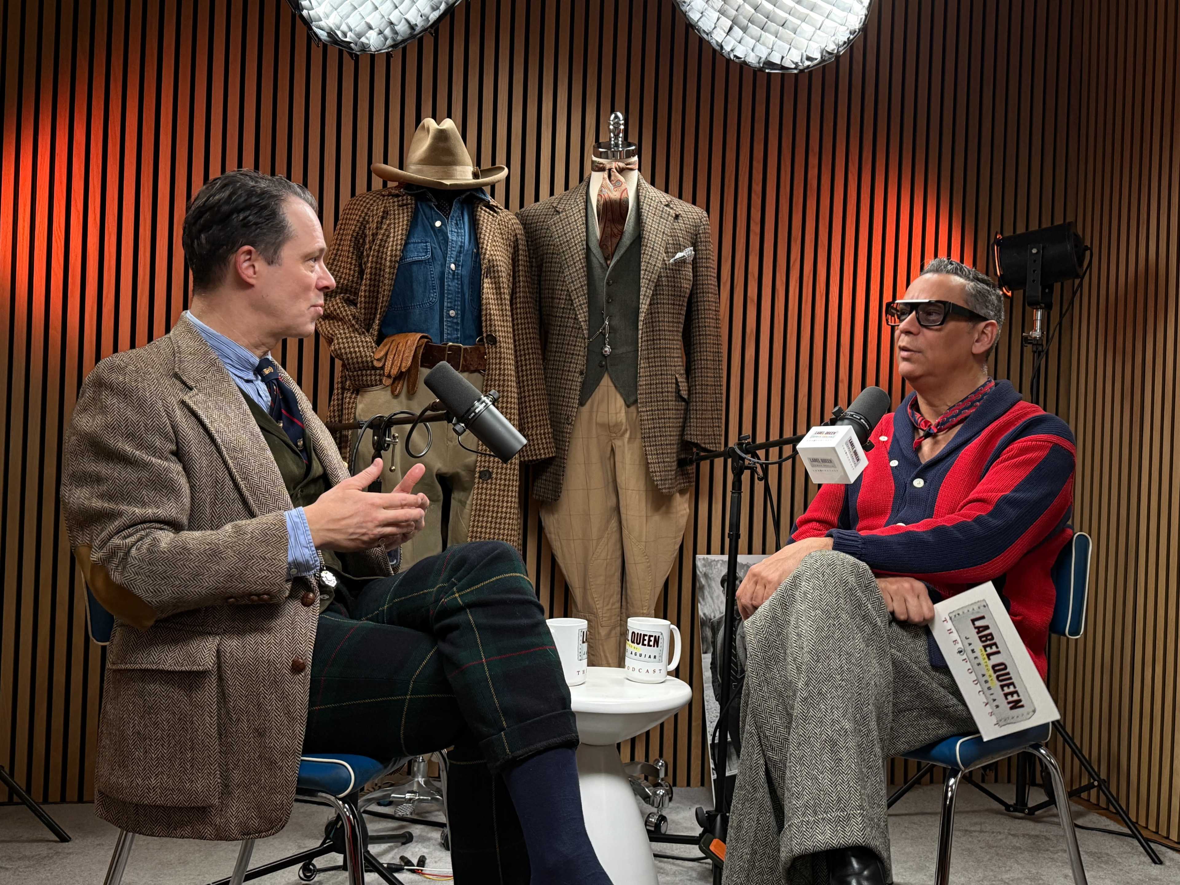 Two men sit in chairs facing each other, engaged in conversation, with stylish clothing displayed on mannequins behind them in a studio setting.