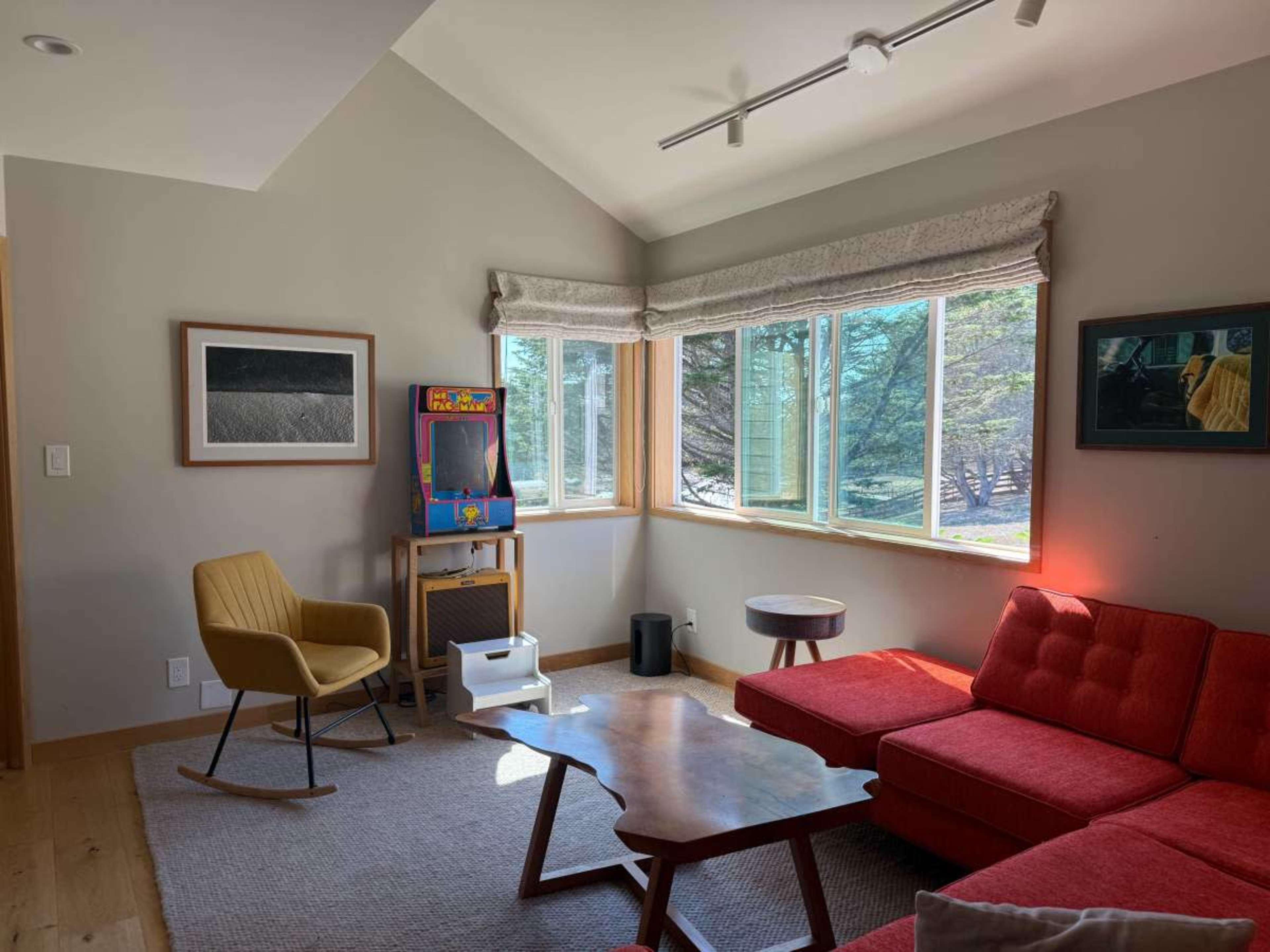 The image shows a living room with a red sectional sofa, a yellow rocking chair, a wooden coffee table, and a vintage arcade game cabinet near large windows.