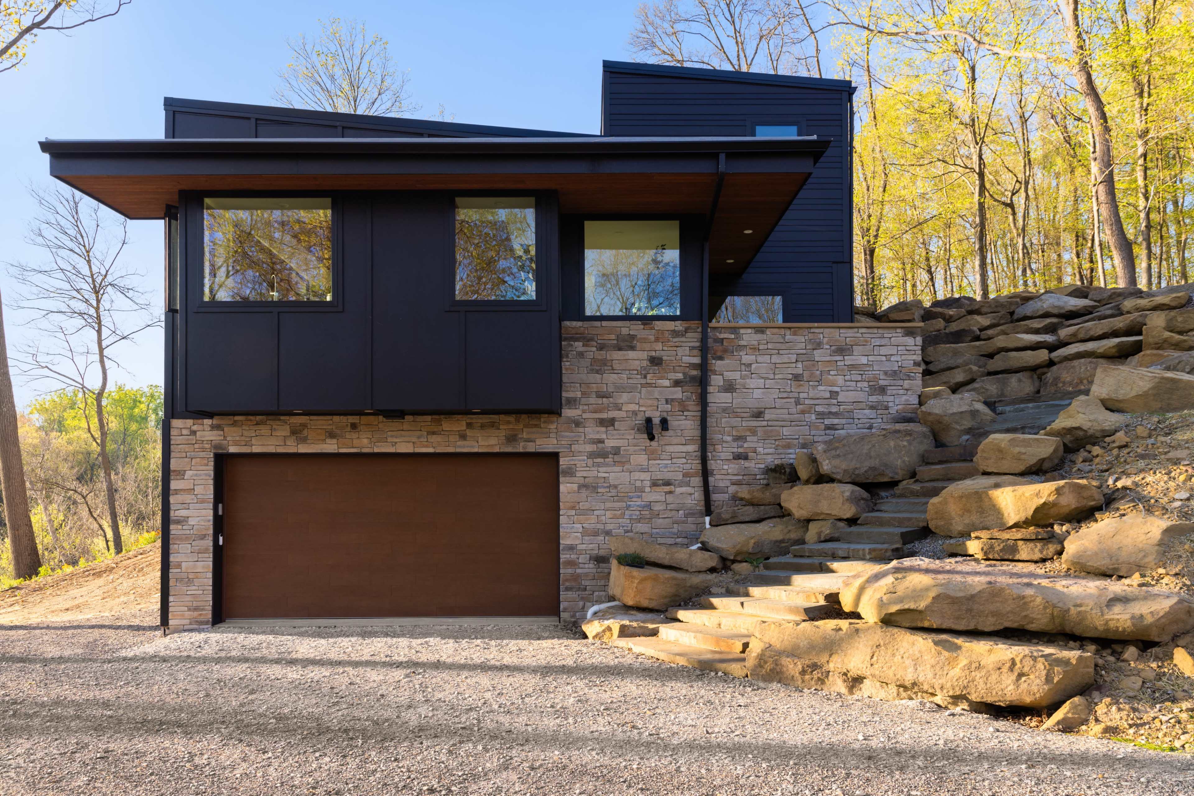 The image shows a modern house with a stone and wood exterior, featuring large windows and a garage, set among rocky landscaping and trees.