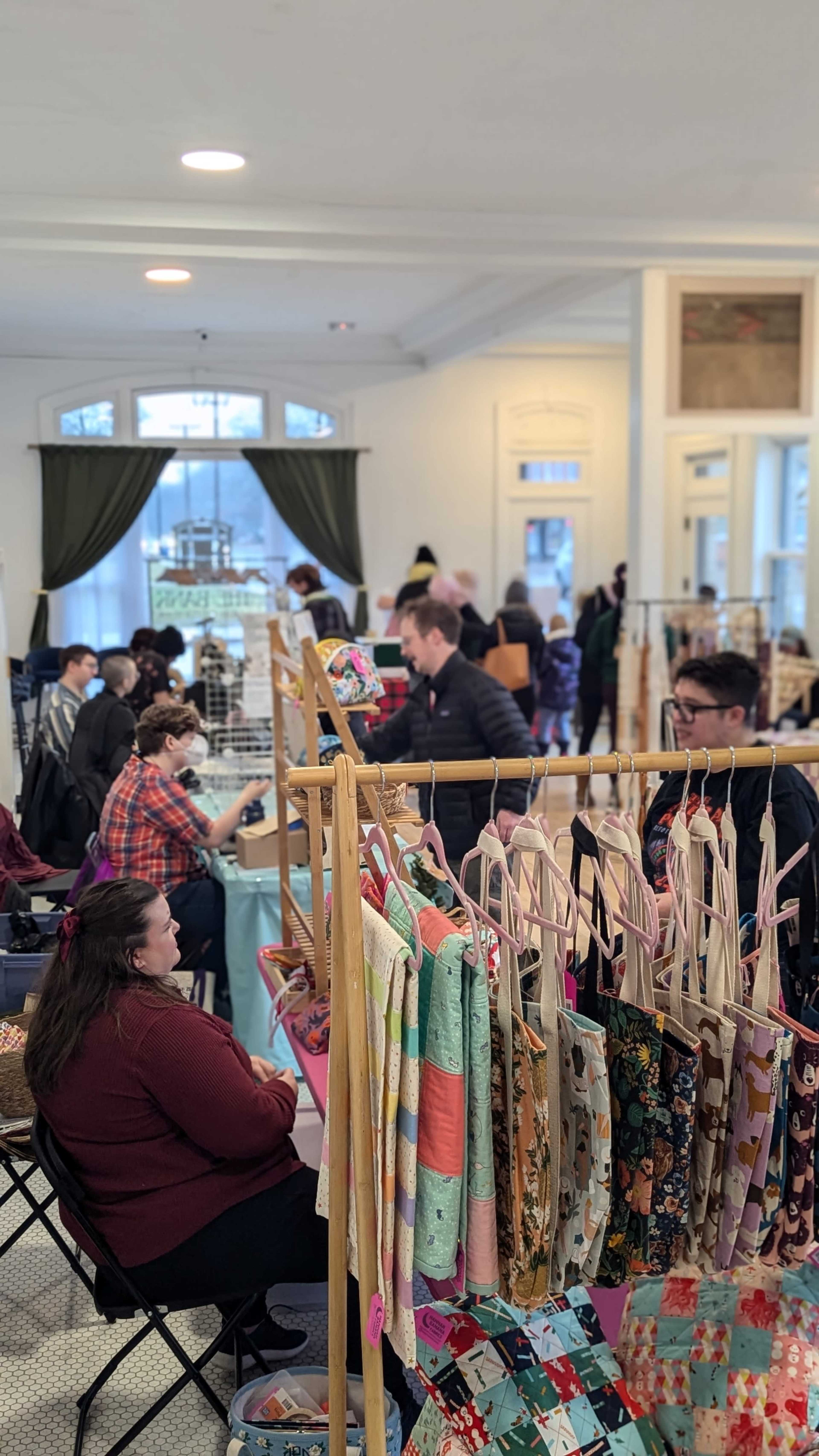 A vibrant market scene features vendors selling handmade items and customers browsing amid tables filled with colorful products.