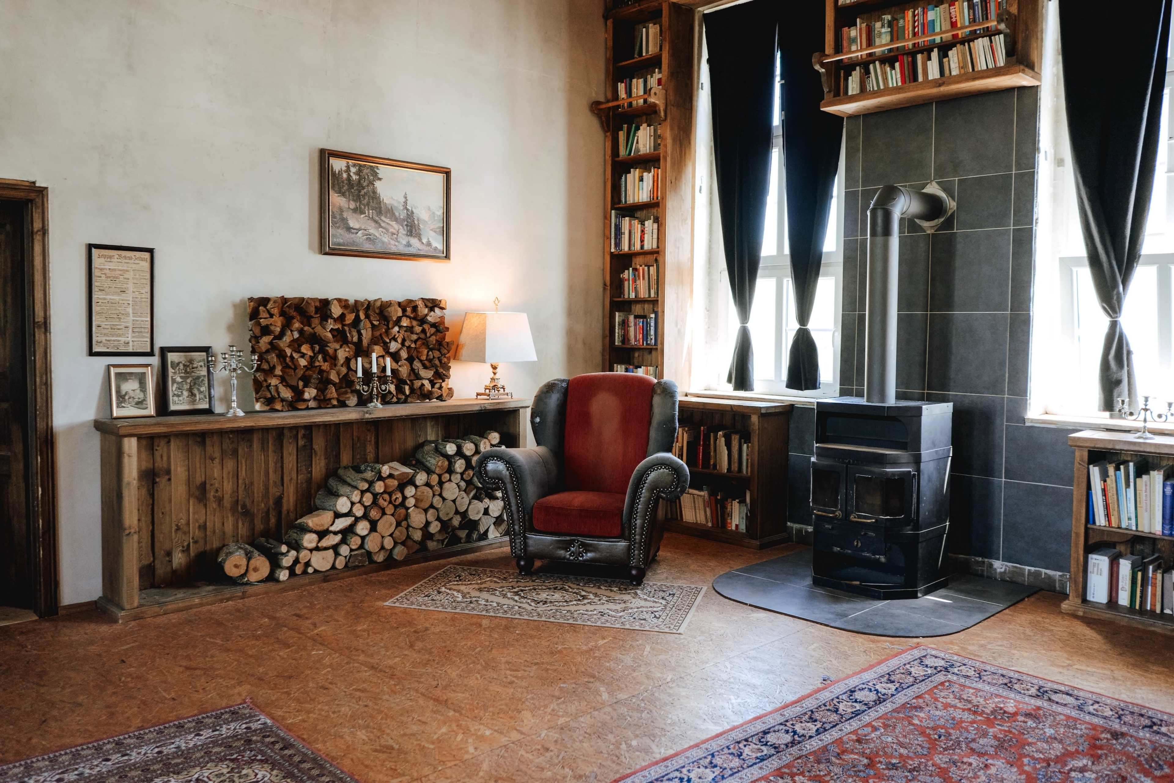 A cozy room features a red armchair beside a wood stove, bookshelves filled with books, and a stack of firewood against the wall.