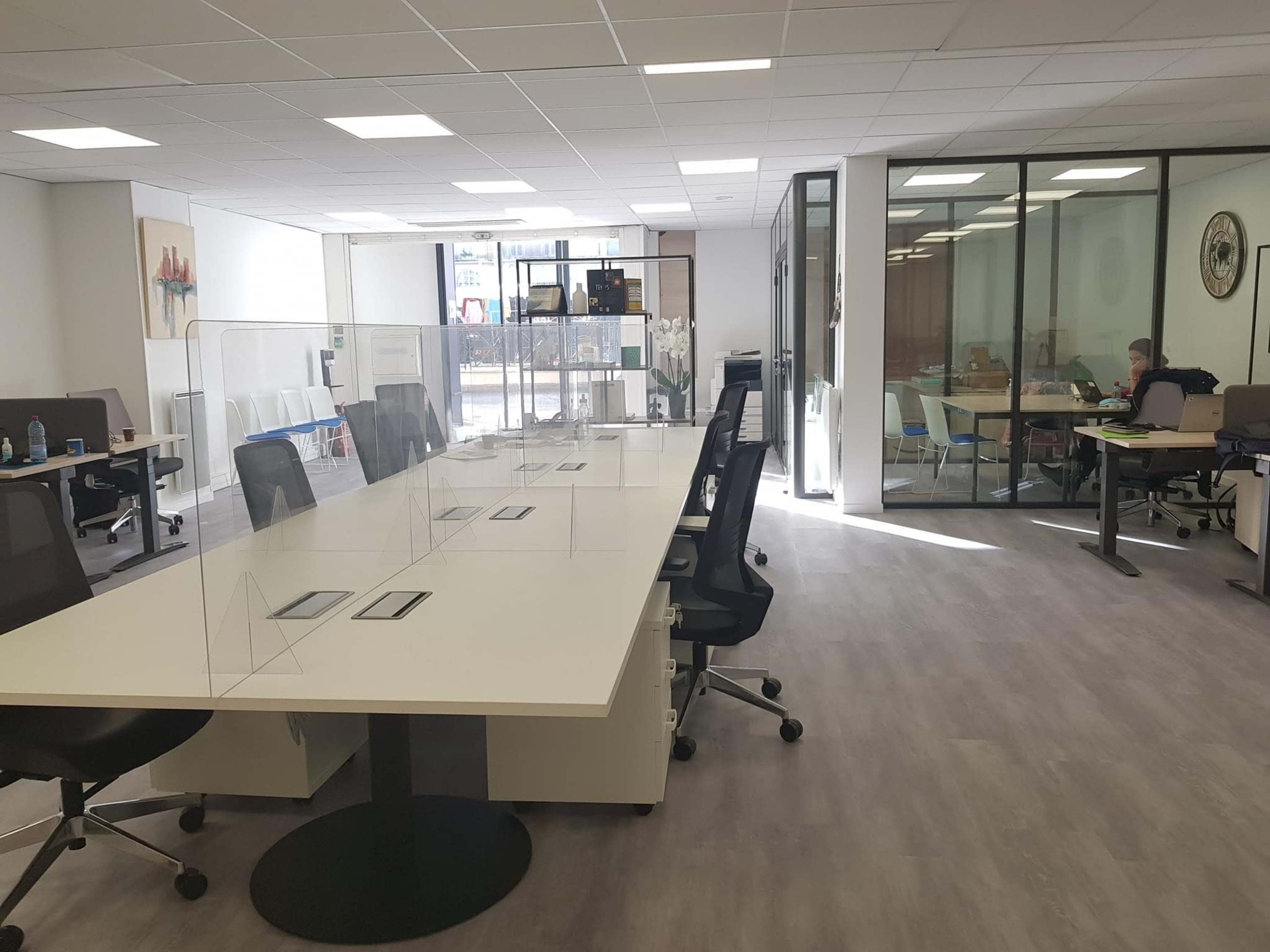 A modern office space with multiple workstations separated by transparent partitions and a glass-walled meeting room in the background.