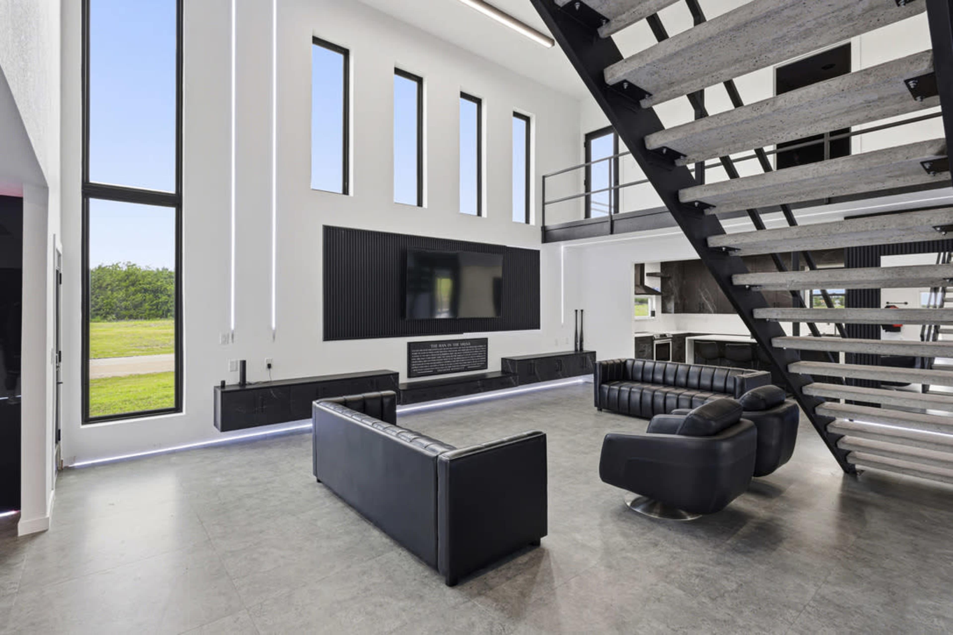 The image shows a modern, spacious living room with large windows, black leather furniture, and a staircase leading to an upper level.
