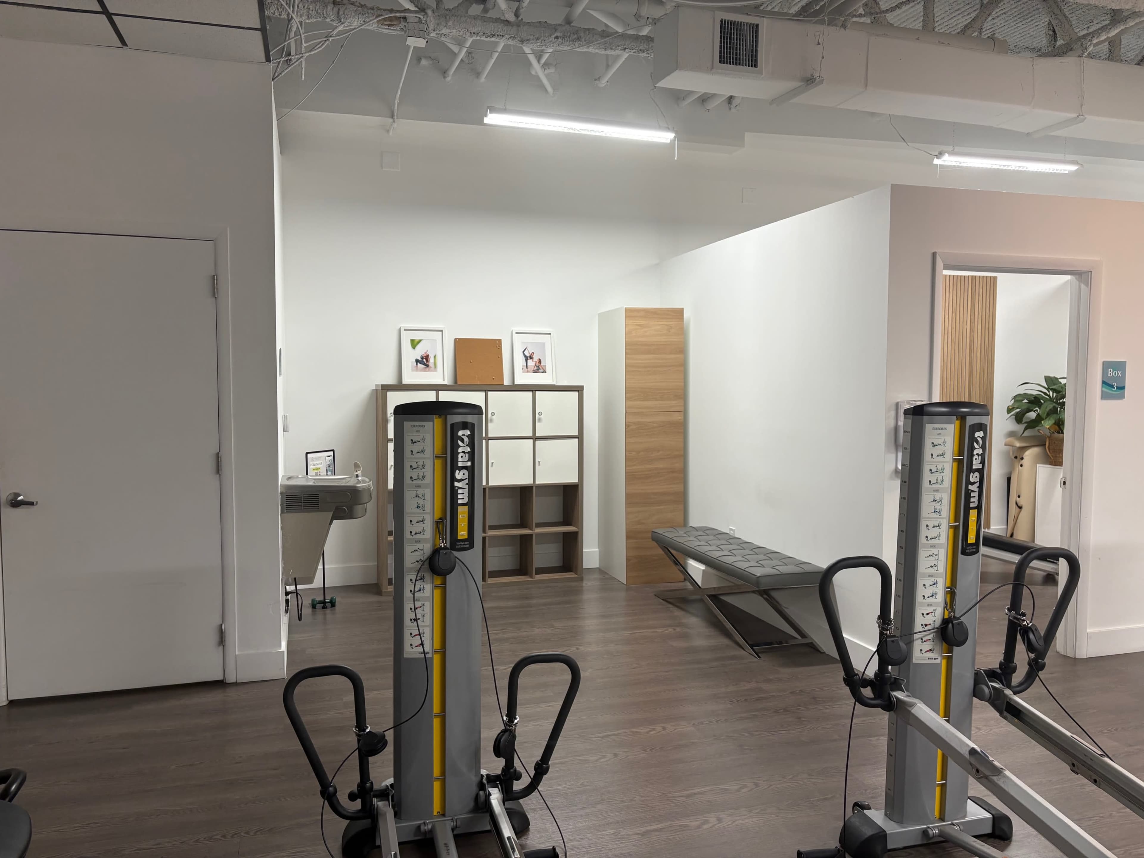 The image shows a fitness area featuring exercise machines, a storage unit, and a wall with framed pictures.