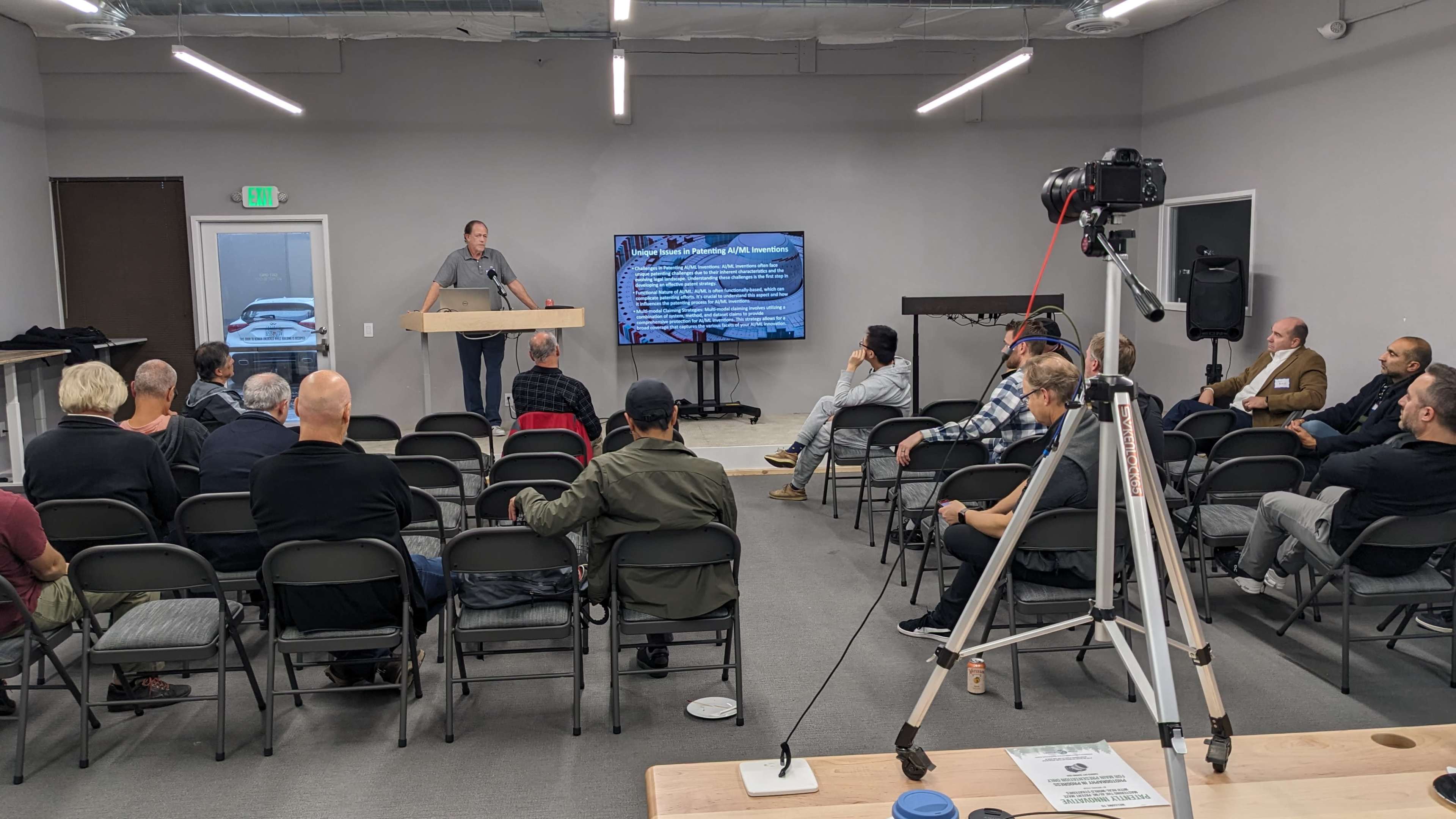 A speaker stands at a podium in front of an audience seated in folding chairs, while a large screen displays information in a gray room.