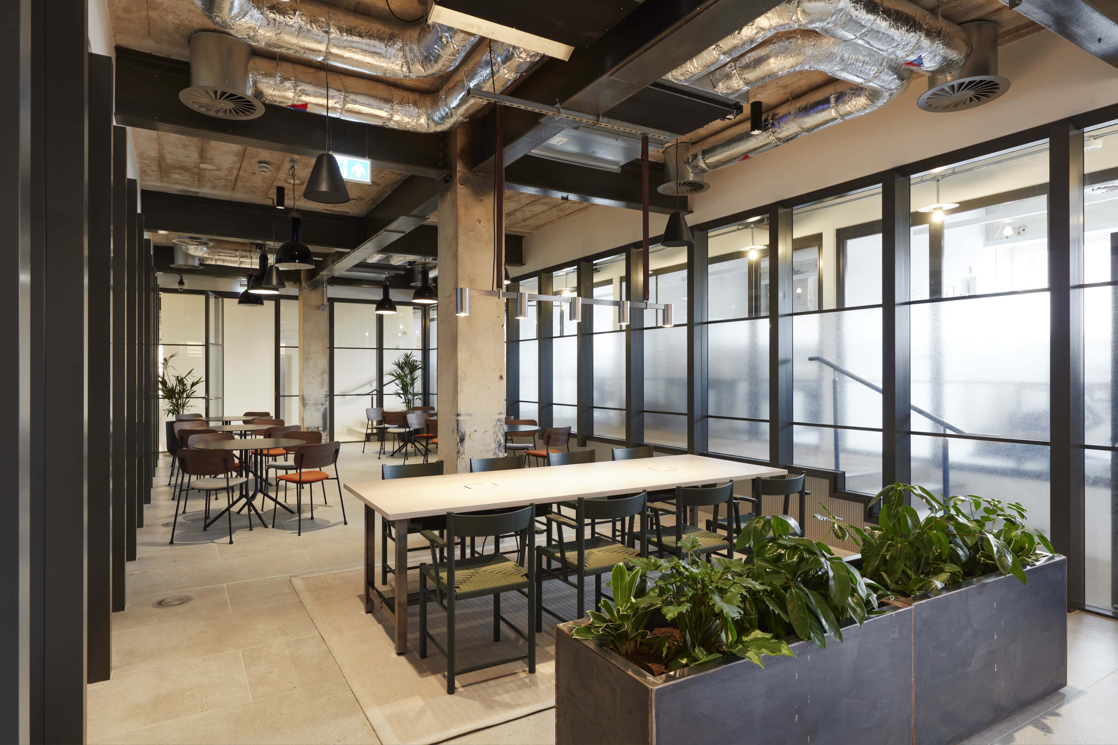 The image shows a modern office space featuring a mix of wooden tables and chairs, large windows, plants, and exposed ductwork on the ceiling.