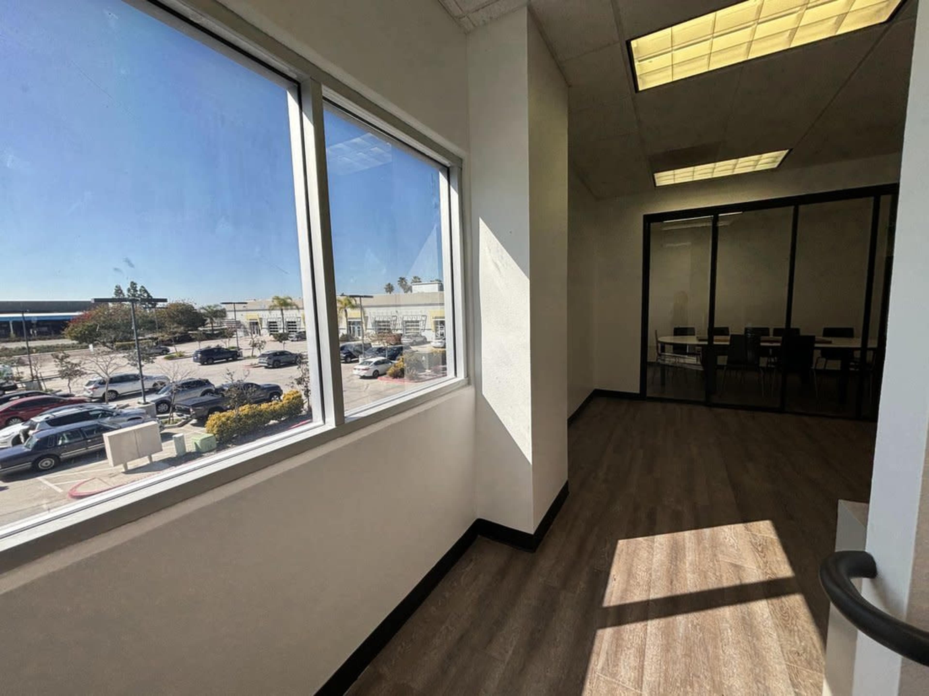 A corner of an office space with large windows overlooking a parking lot and a meeting room in the background.
