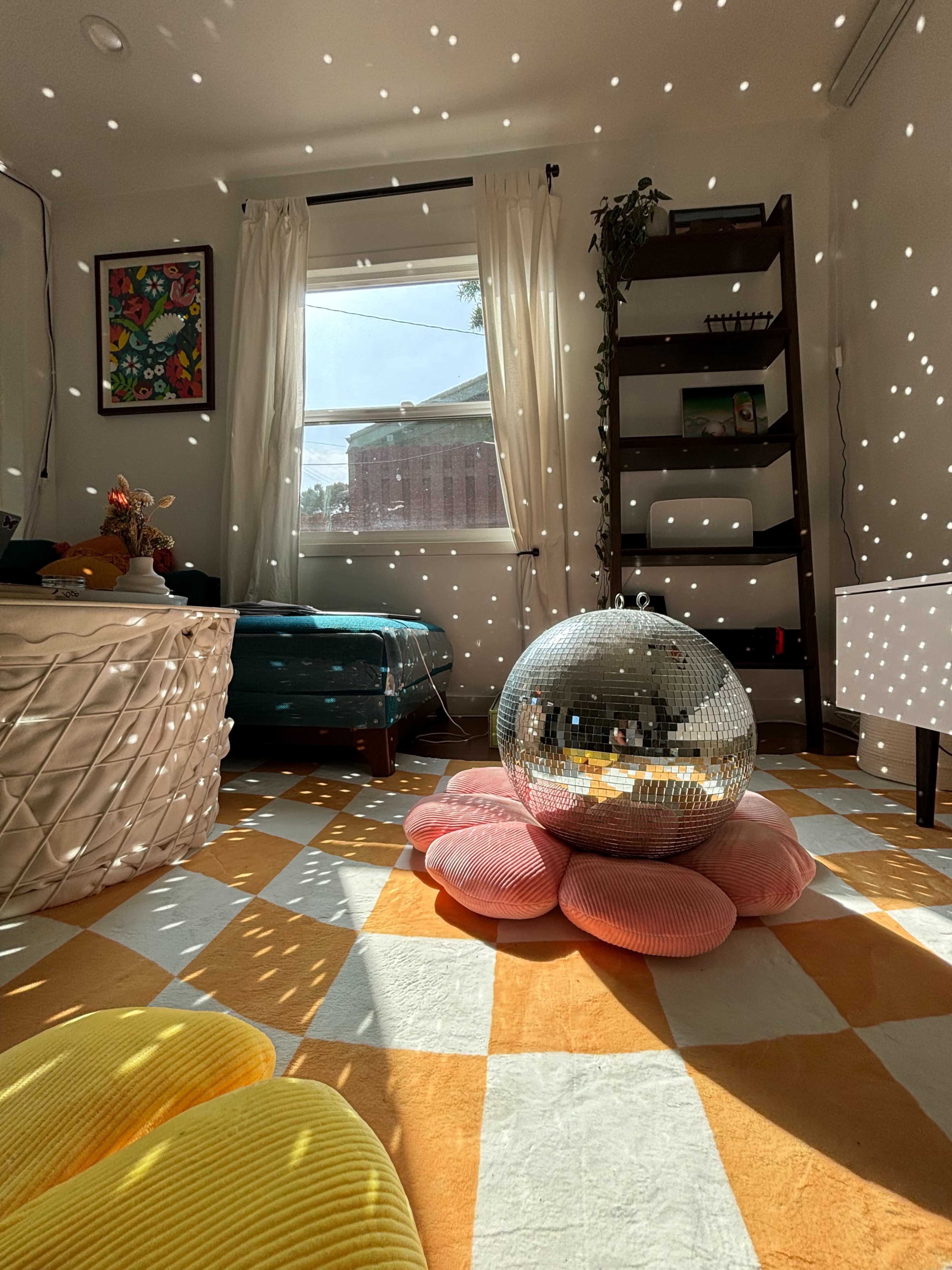 The room features a disco ball on a pink flower-shaped rug, surrounded by colorful cushions and illuminated by sunlight filtering through a window, casting scattered light patterns on the walls.