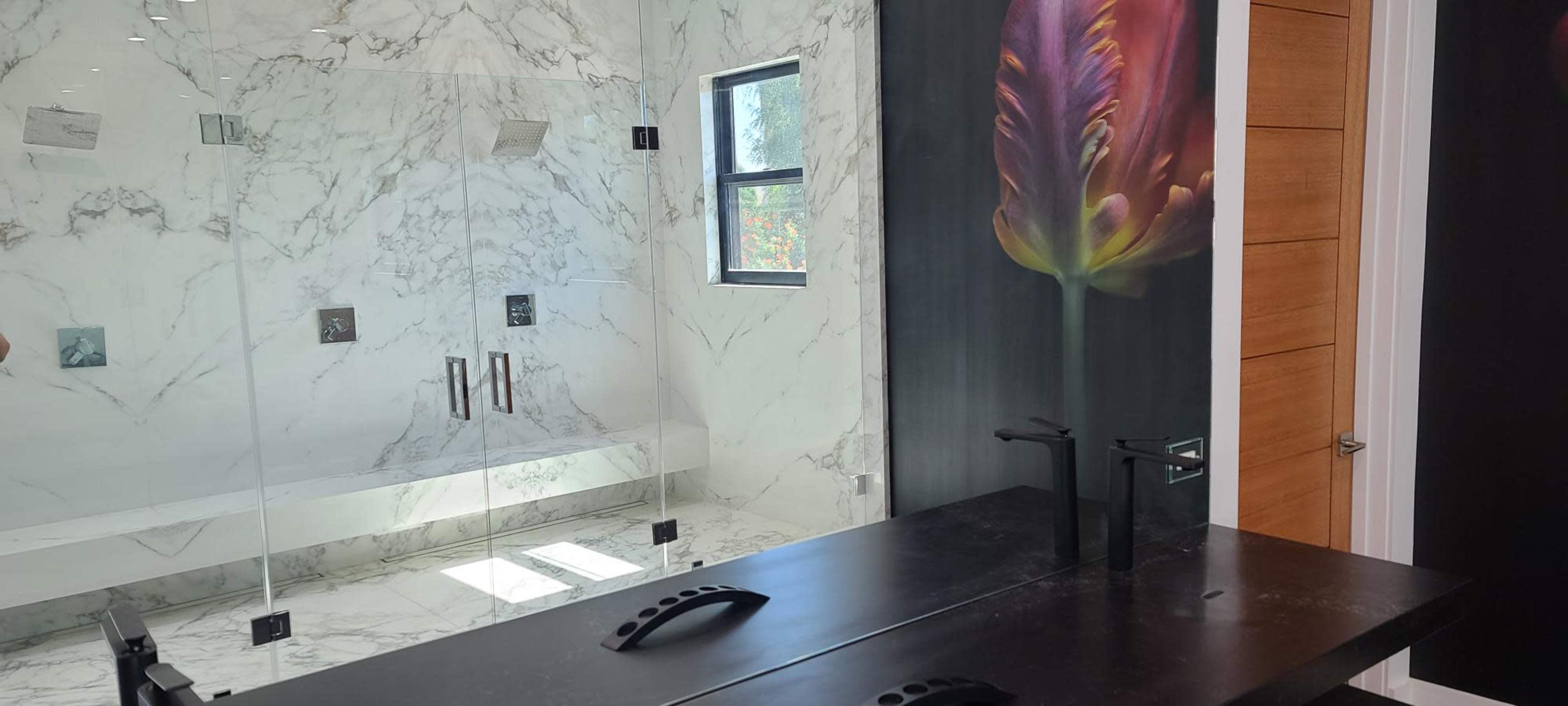 A modern bathroom featuring a glass shower, marble walls, and a dark countertop with dual faucets.