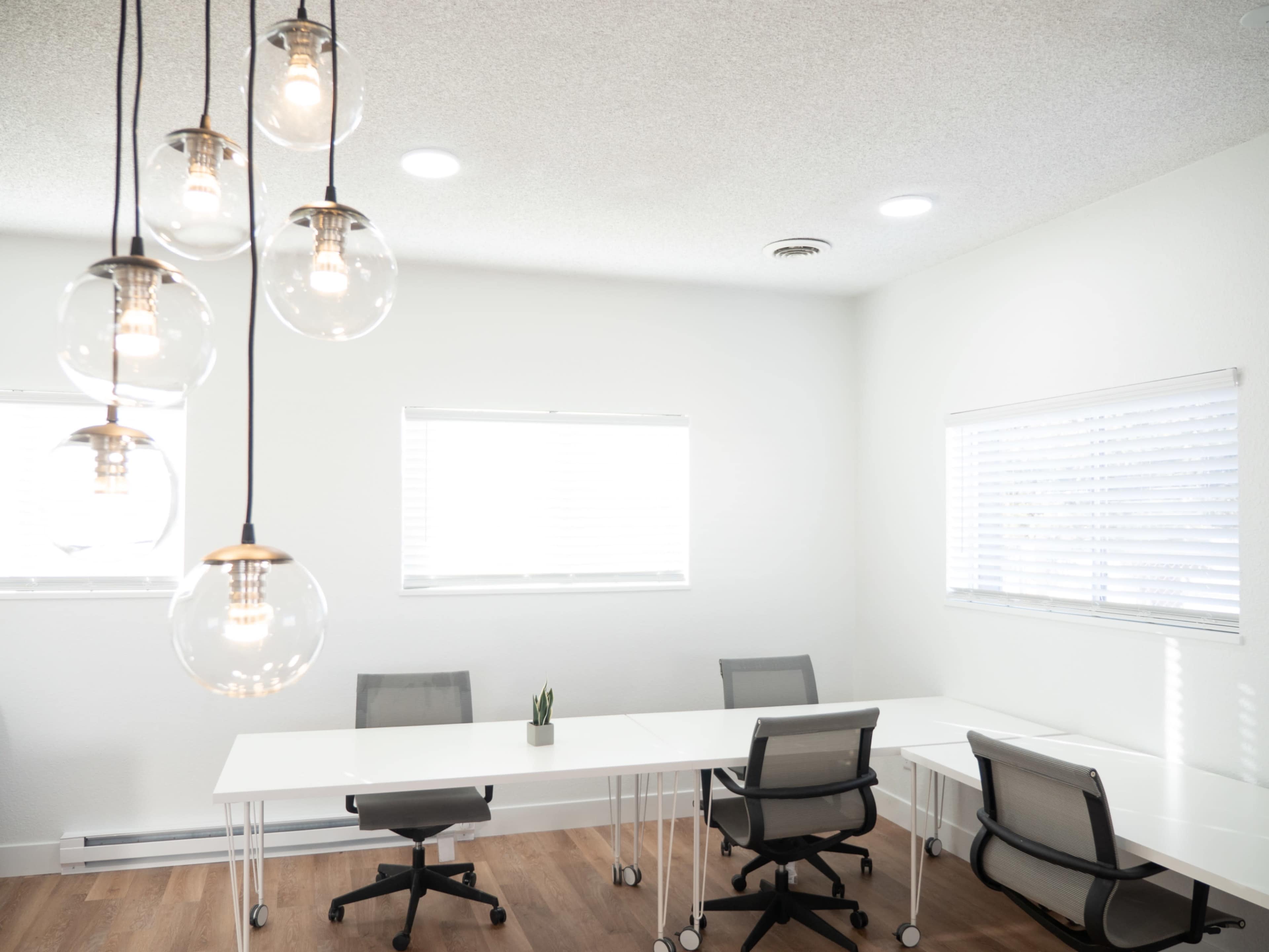 A modern office space features two large tables with rolling chairs and light fixtures hanging from the ceiling.