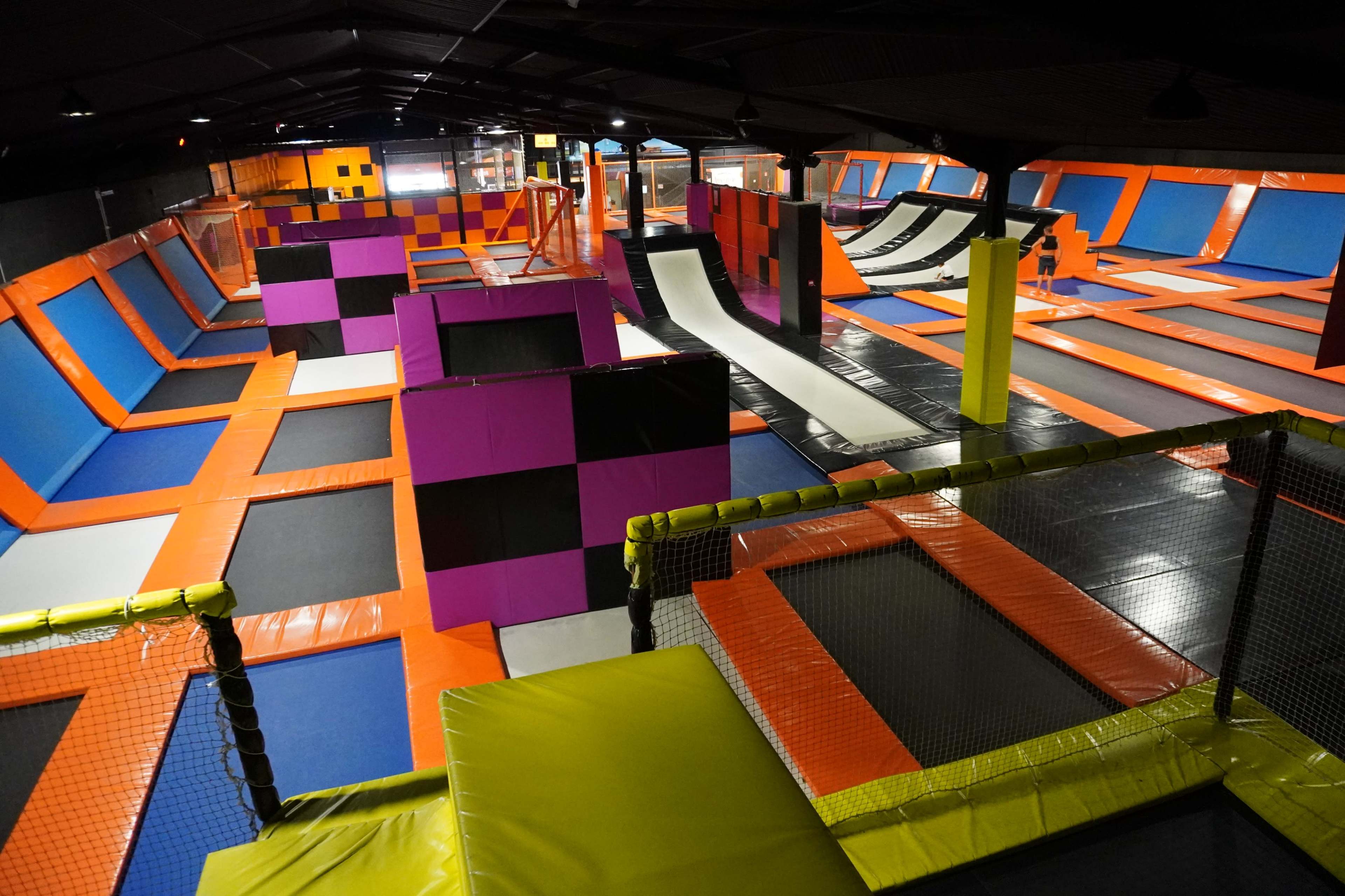 A large indoor trampoline park featuring multiple interconnected trampolines, towering foam pit structures, and various angled surfaces for jumping and play.