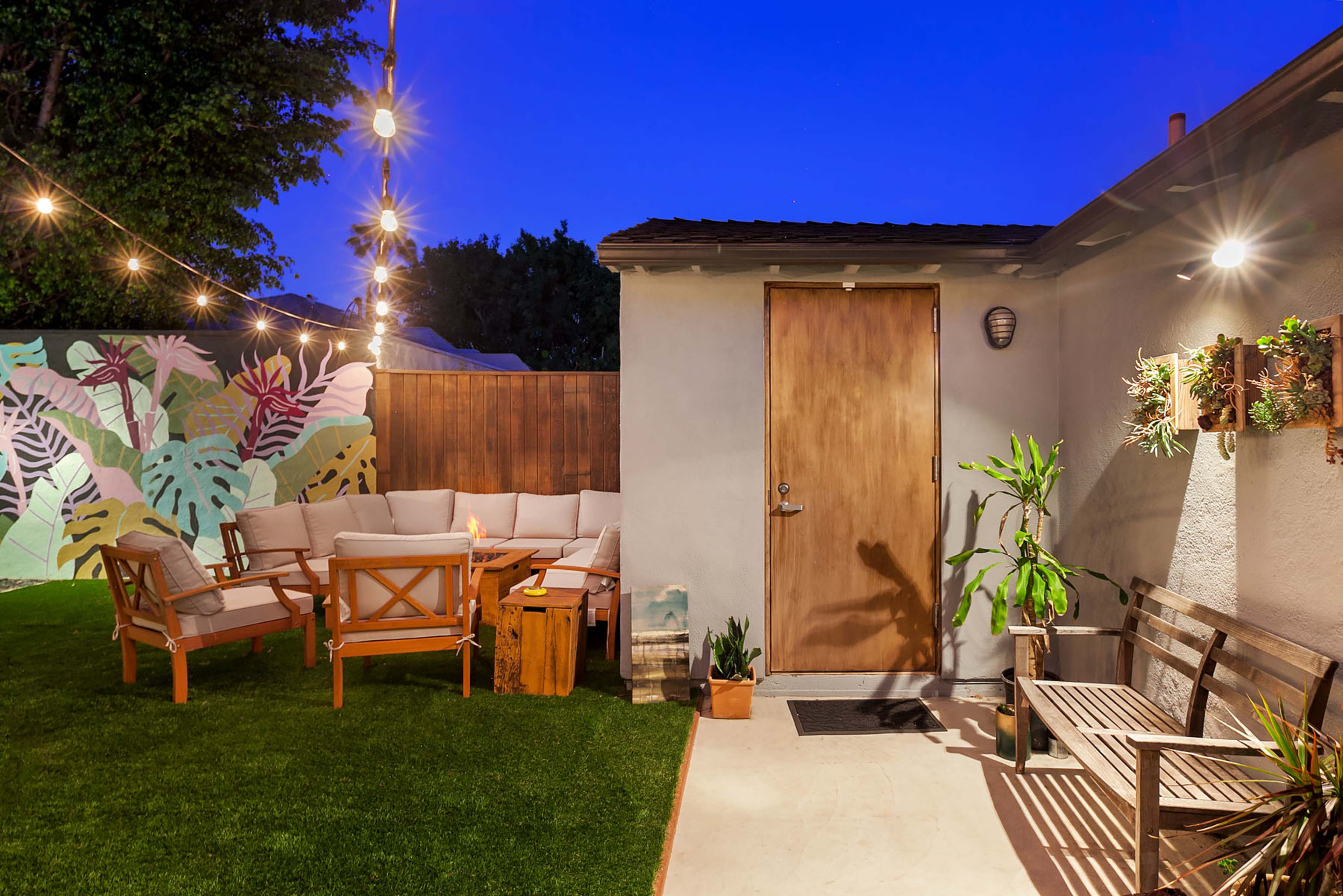 A backyard area featuring a wooden door, outdoor seating arrangements, decorative plants, and string lights against a twilight sky.