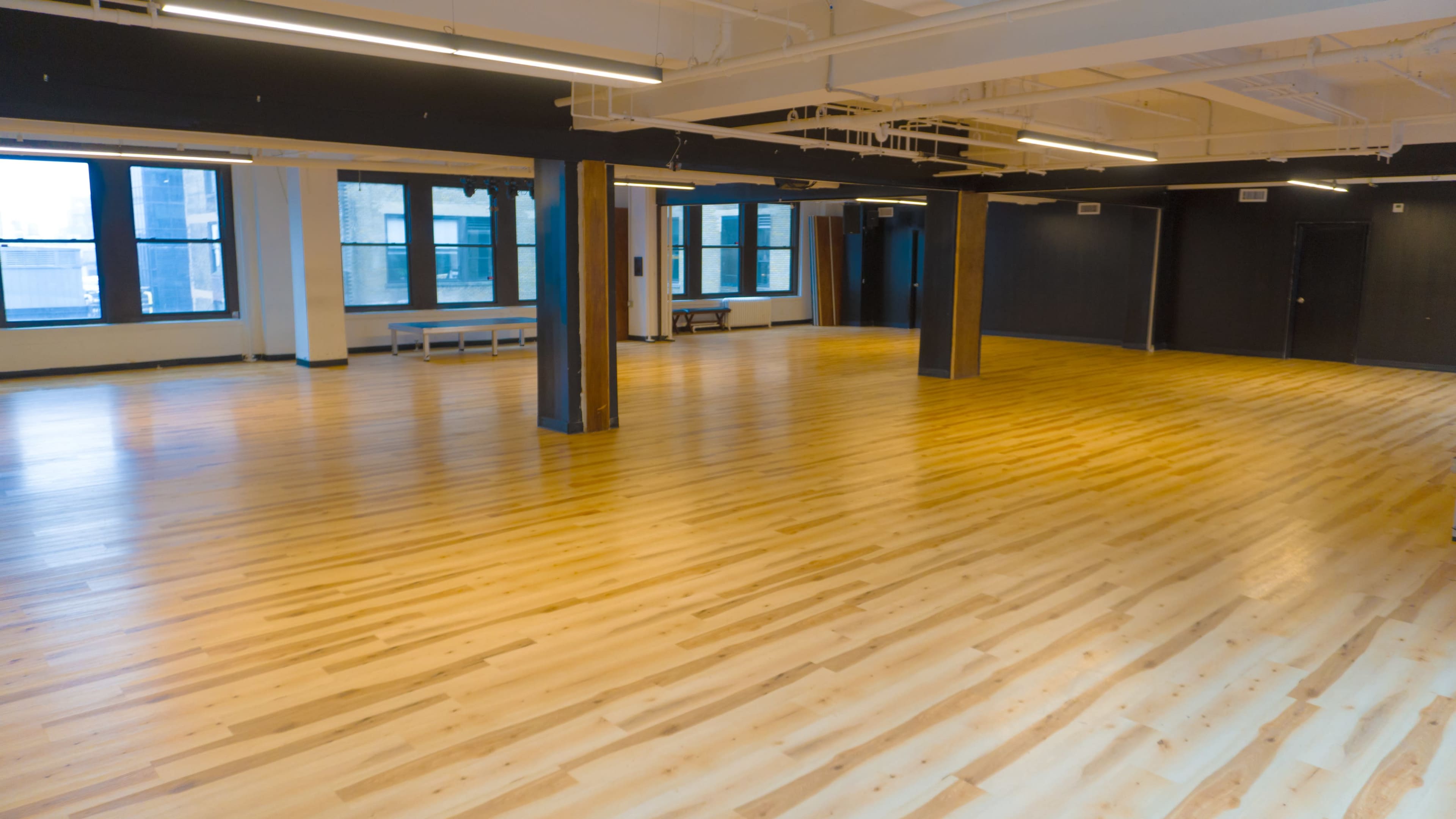 The image shows a spacious, empty room with polished wood flooring and large windows allowing natural light to enter.