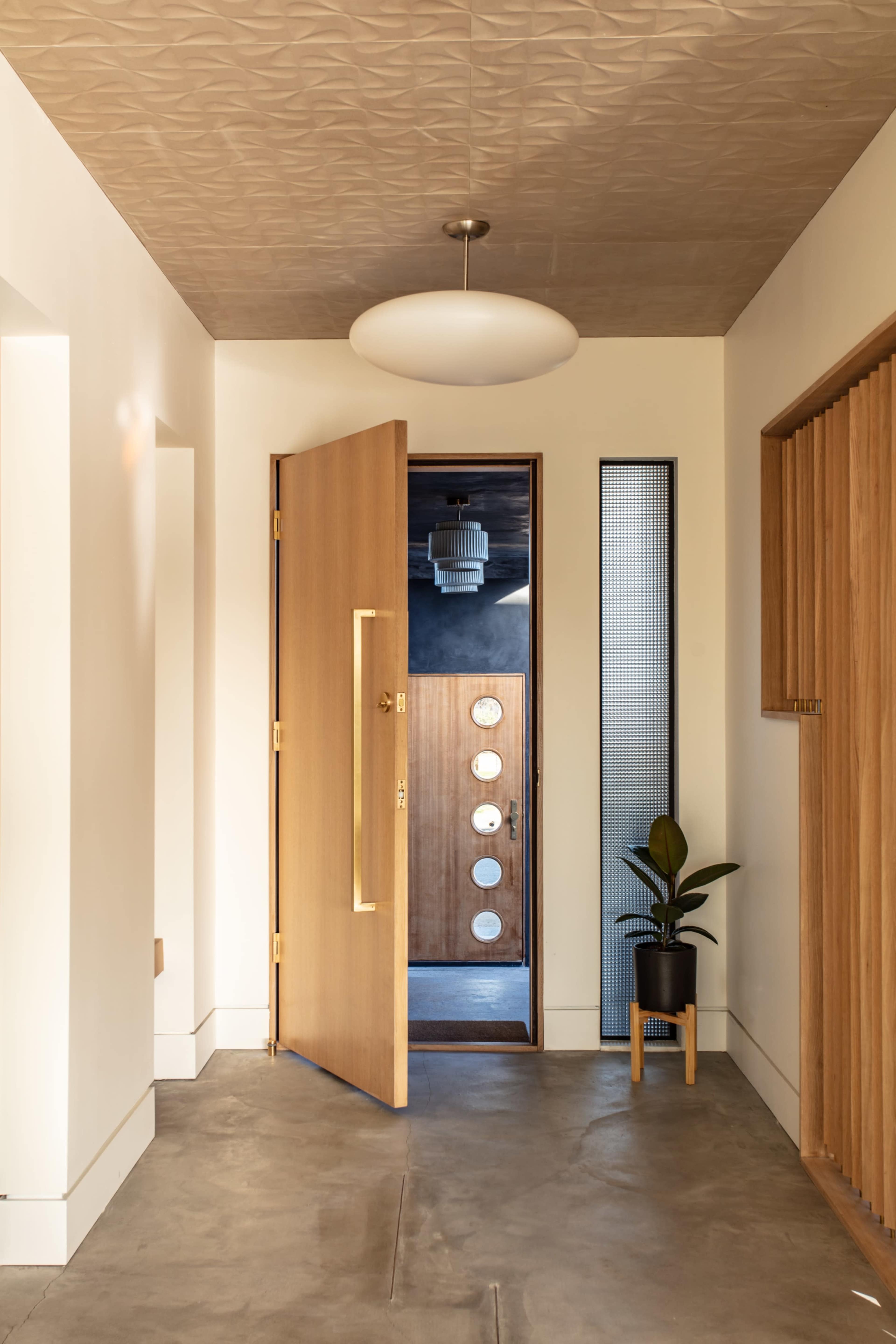 A wooden door stands open, revealing a modern interior with a potted plant and textured ceiling.