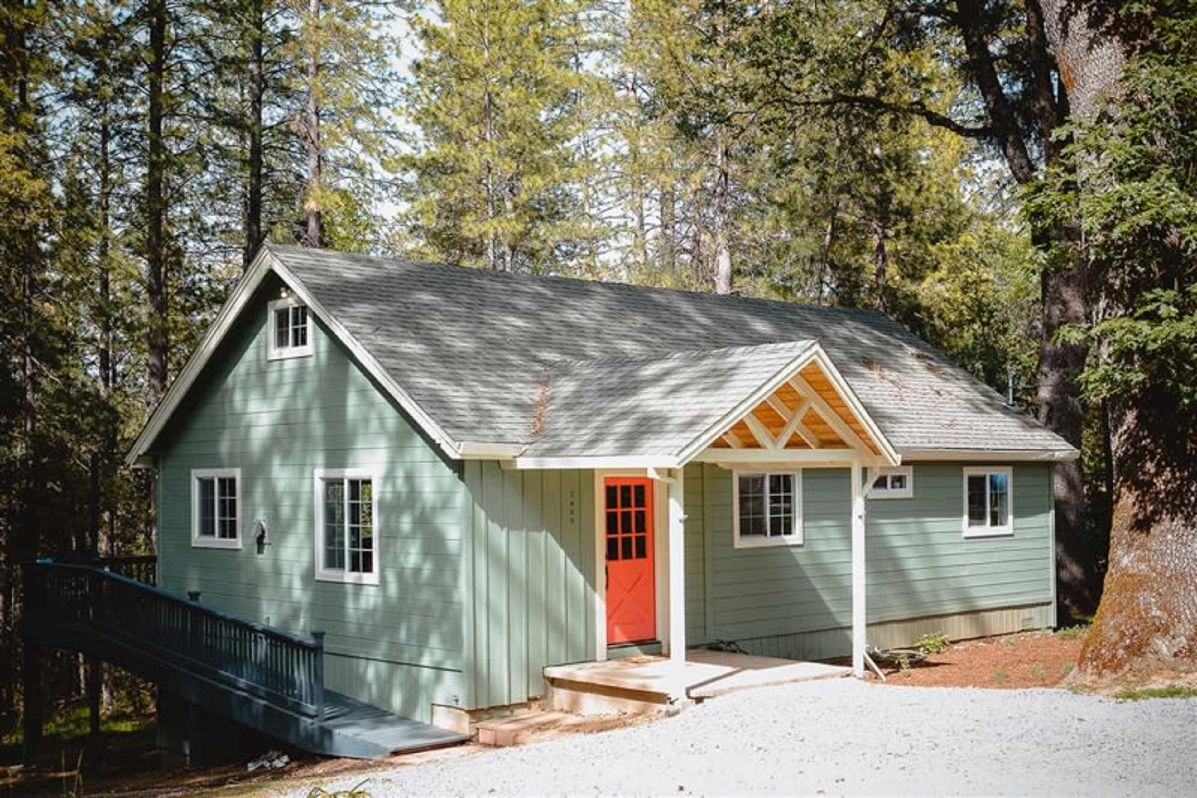 A light green house with a red front door, surrounded by tall trees and a gravel path in a forested area.