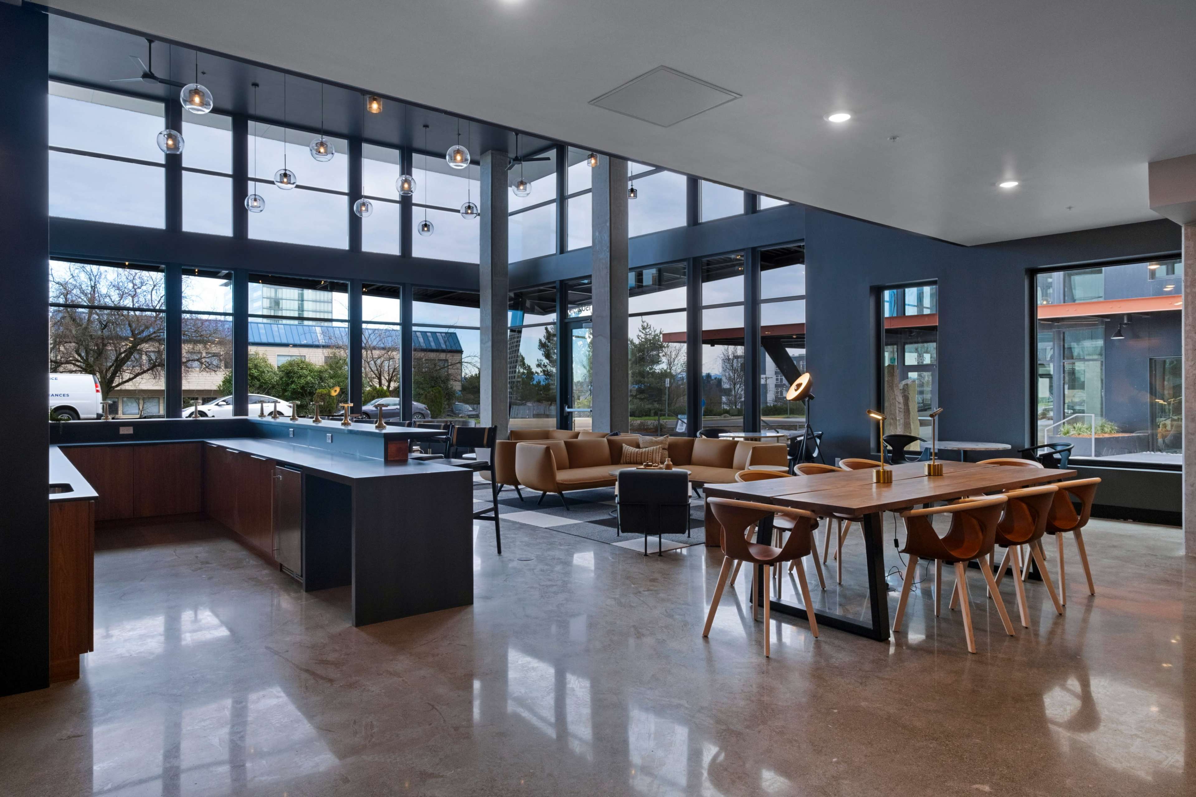 The image shows a modern interior space featuring a kitchen area, dining table, and lounge area with large windows allowing natural light.