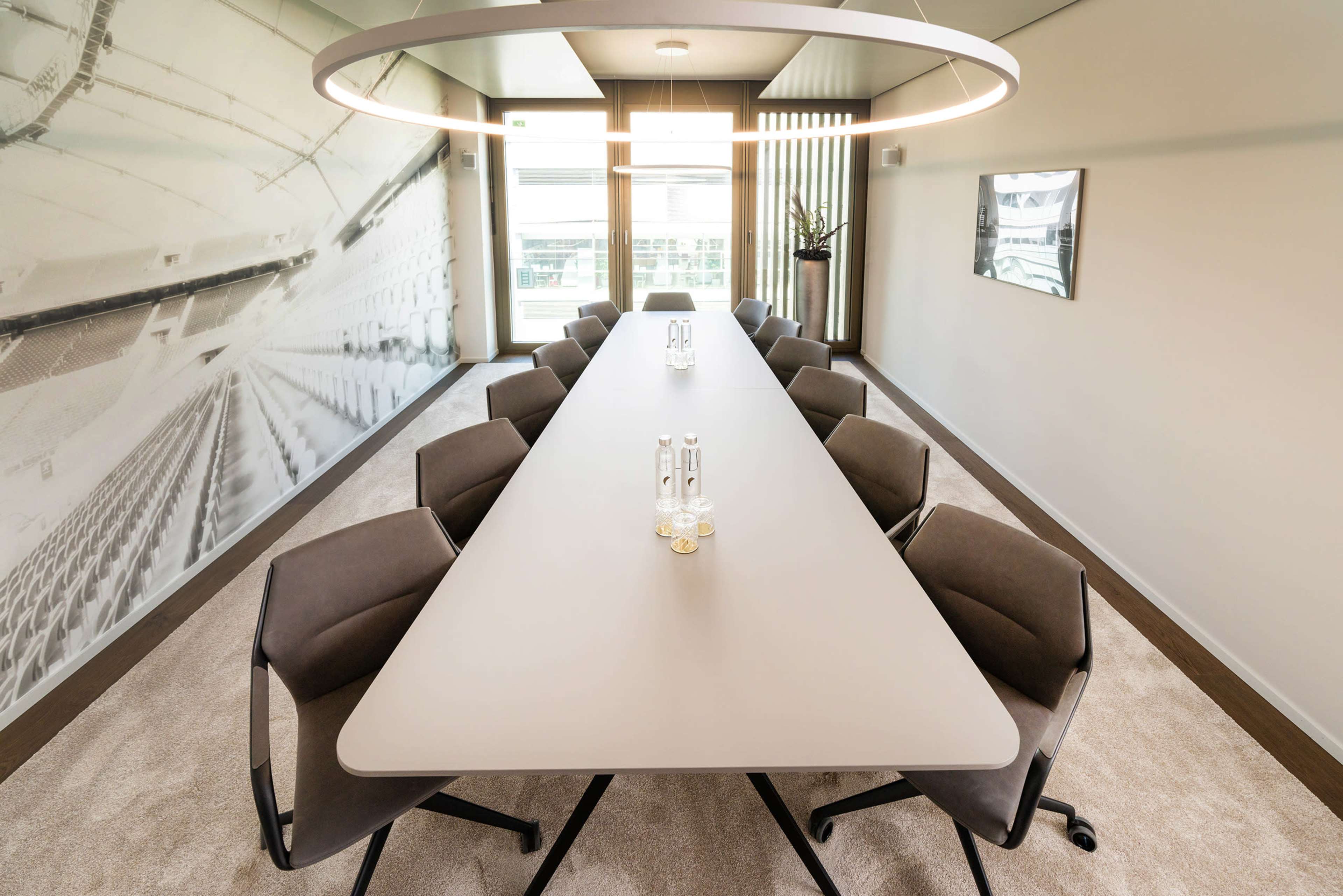 The image shows a modern conference room with a long, triangular table surrounded by ergonomic chairs and a circular light fixture overhead.