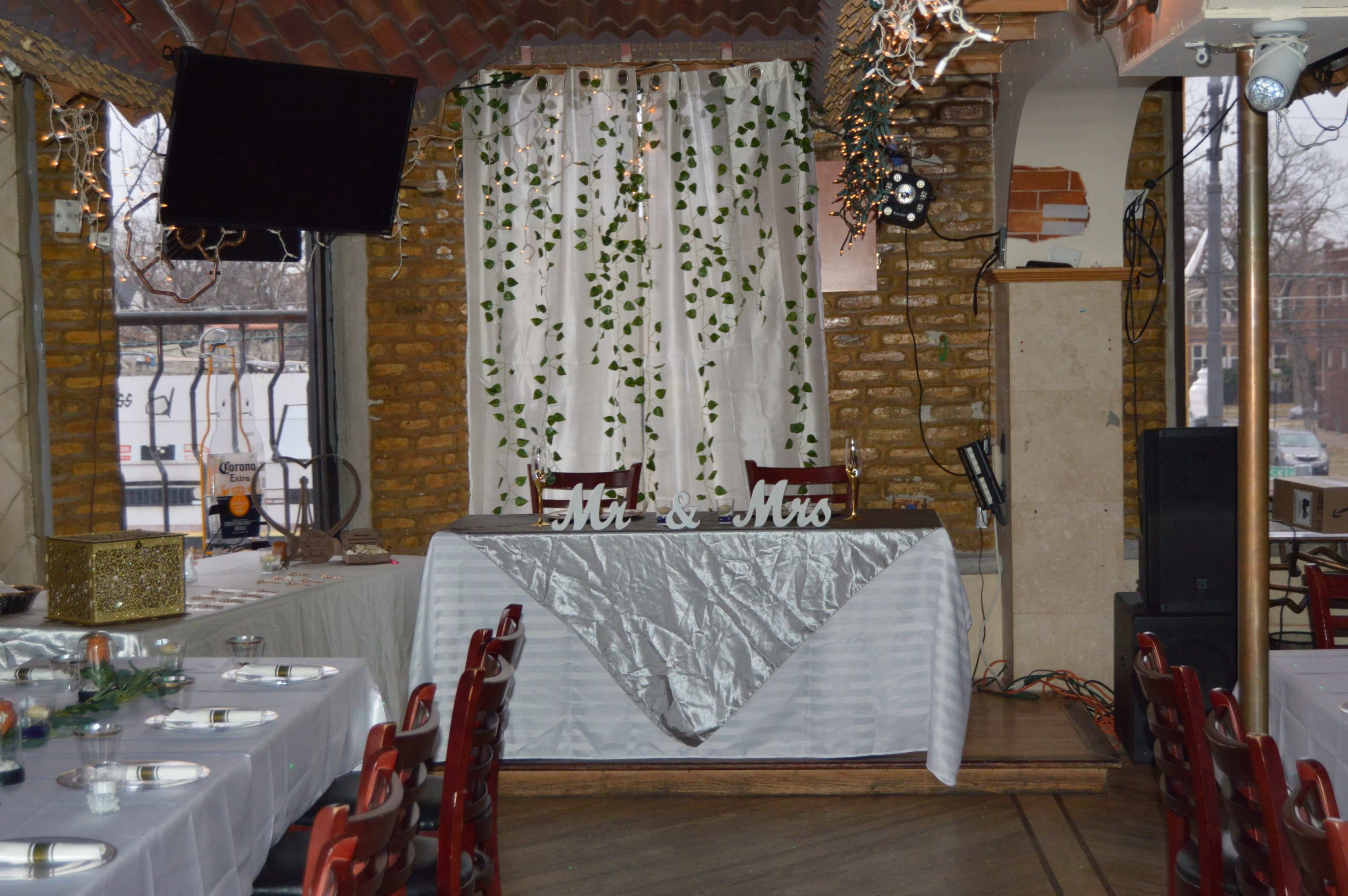 A decorated table with a white tablecloth and a "Mr. & Mrs." sign is set up in a venue, surrounded by festive decor and draped curtains.