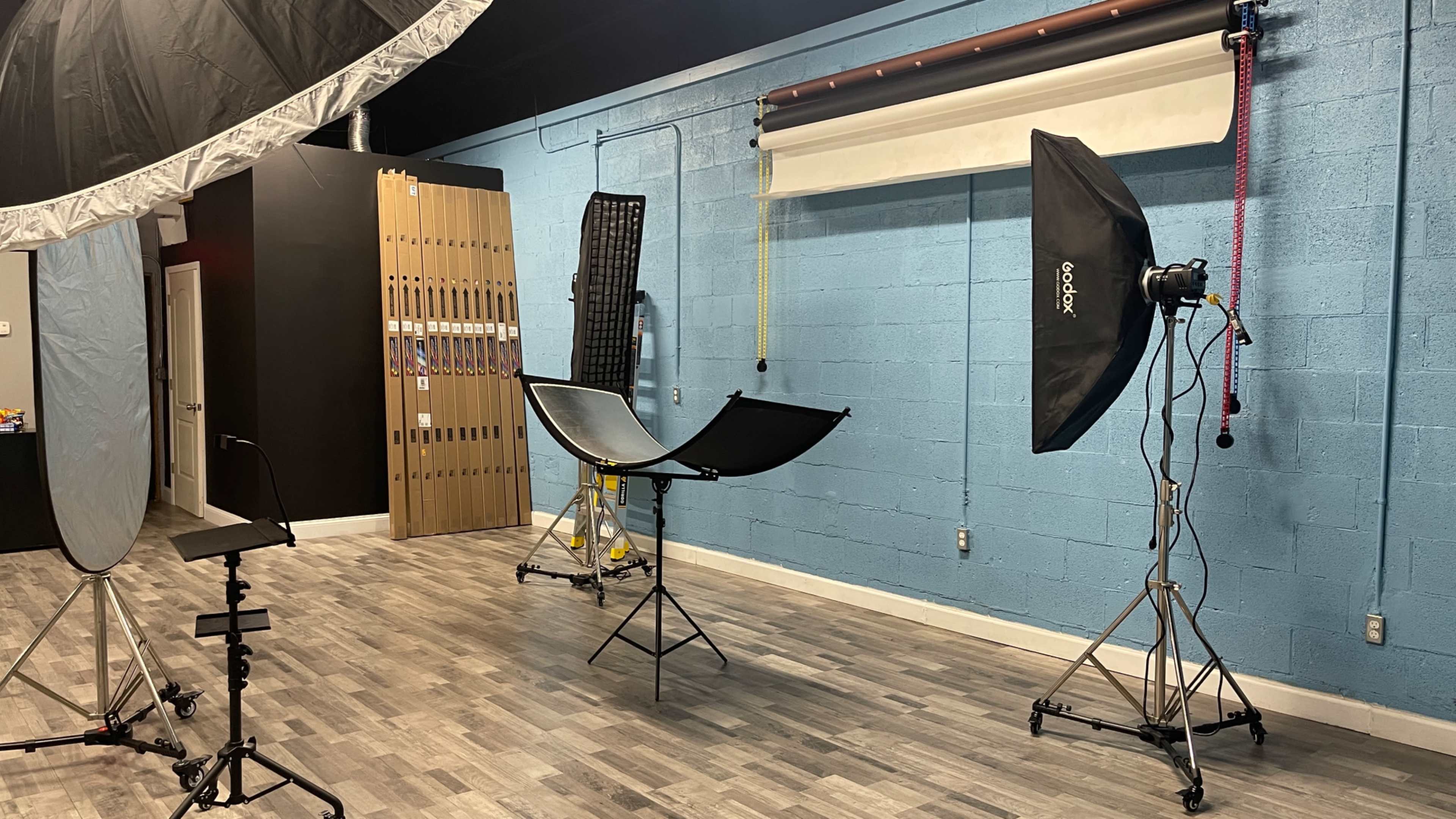 The image shows a photography studio featuring multiple lighting equipment setups, including softboxes and a reflector, against a blue wall.